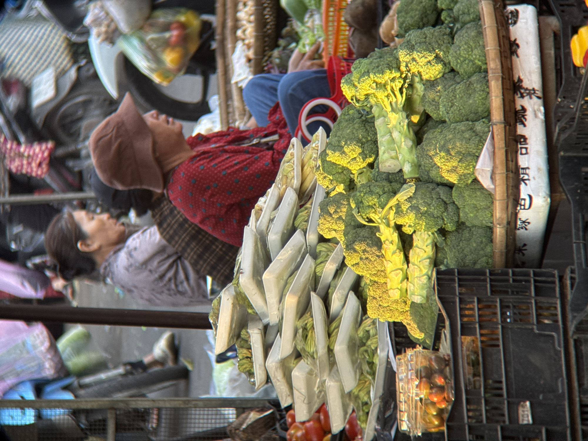 In Nha Trang, Vietnam, a street market scene unfolds with a variety of fresh produce and vendors. In the foreground, a pyramid of vegetables, primarily broccoli and what appear to be string beans or asparagus, is displayed in white styrofoam trays stacked on a dark crate. To the left, a clear plastic container holds small, dark red tomatoes or berries. Behind the produce, a woman wearing a brown hat and a red, polka-dotted shirt is seated, partially visible. She is looking to her right. Another woman, seen from the side and slightly blurred, is wearing a light purple top and has dark hair. She is also looking to her right. In the background, motorcycles and more market stalls are visible, suggesting a bustling marketplace. The lighting indicates it is daytime, with sunlight casting shadows and highlighting the textures of the vegetables. The overall impression is one of a vibrant and active local food market.