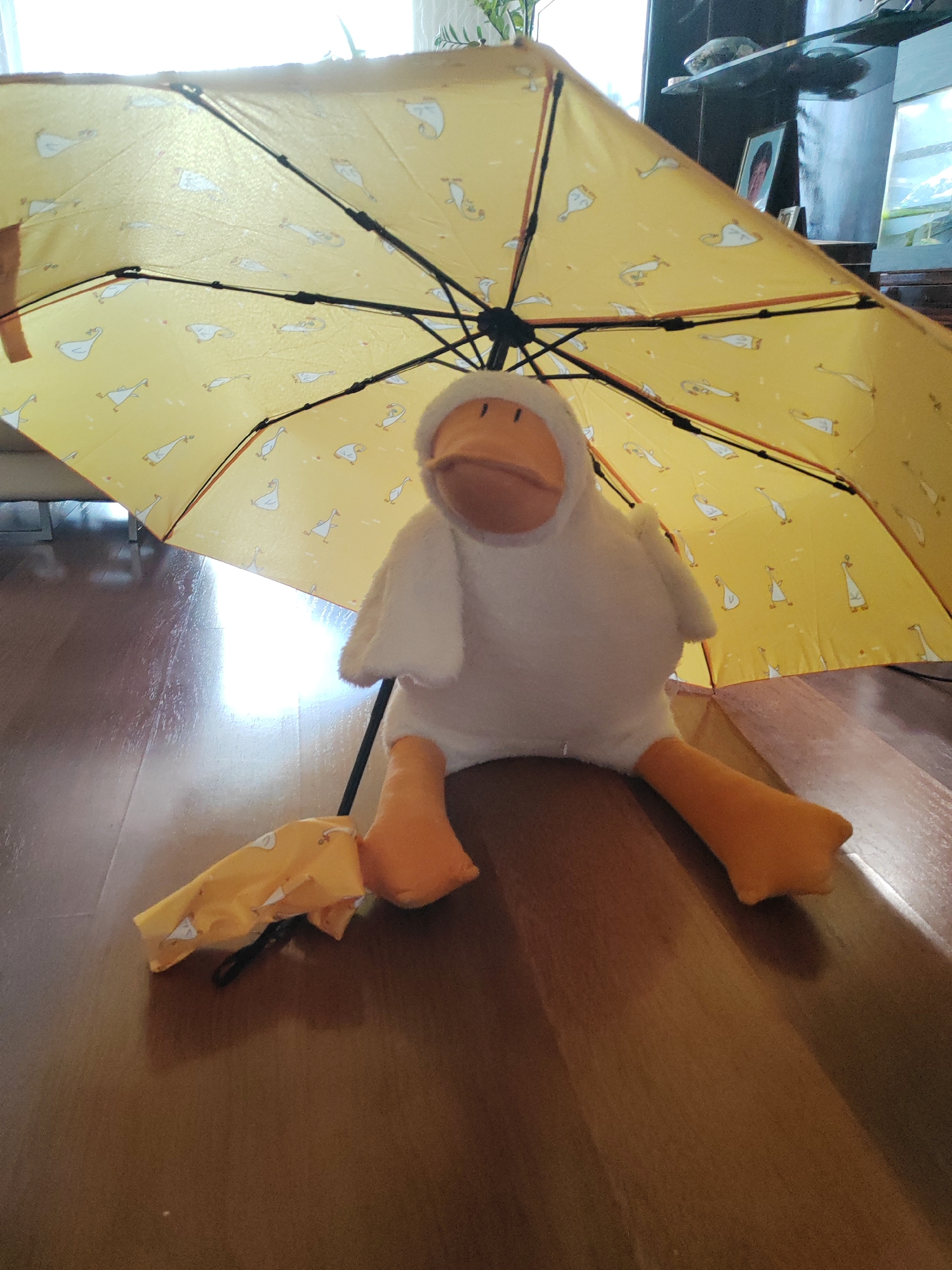 A plush toy resembling a duck, wearing a white hooded outfit, sits on a polished wooden floor beneath an open yellow umbrella adorned with a pattern of white ducks. The umbrella's internal structure of black metal spokes is visible. To the left of the duck toy, a folded yellow fabric with a similar duck pattern and a black strap lies on the floor. The background shows a shelf with decorative items, including framed pictures, and a window with white curtains allowing light to filter in. The lighting suggests it is daytime. The scene appears to be indoors. This image does not provide any information that would allow it to be associated with the city of Kemerovo, Russia.