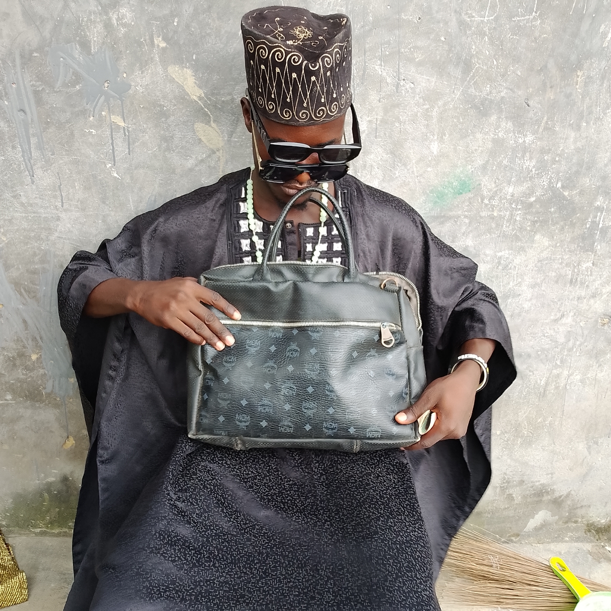 A medium shot captures a Black person seated against a rough, grey concrete wall in Ado, Nigeria. The person wears a traditional black garment with a patterned fabric and a black, ornate cap adorned with gold embroidery. Dark sunglasses are perched on their nose, and two strands of light green beaded necklaces hang around their neck. They are holding a black, textured leather handbag with multiple zippered compartments and a prominent MCM logo pattern. The person's right hand rests on the bag, while their left hand grips its handle. A bright yellow and green dustpan and a broom made of natural fibers are visible on the lower right side of the image, suggesting an outdoor or semi-outdoor setting with some cleaning equipment present. The overall scene is static and appears to be a portrait.