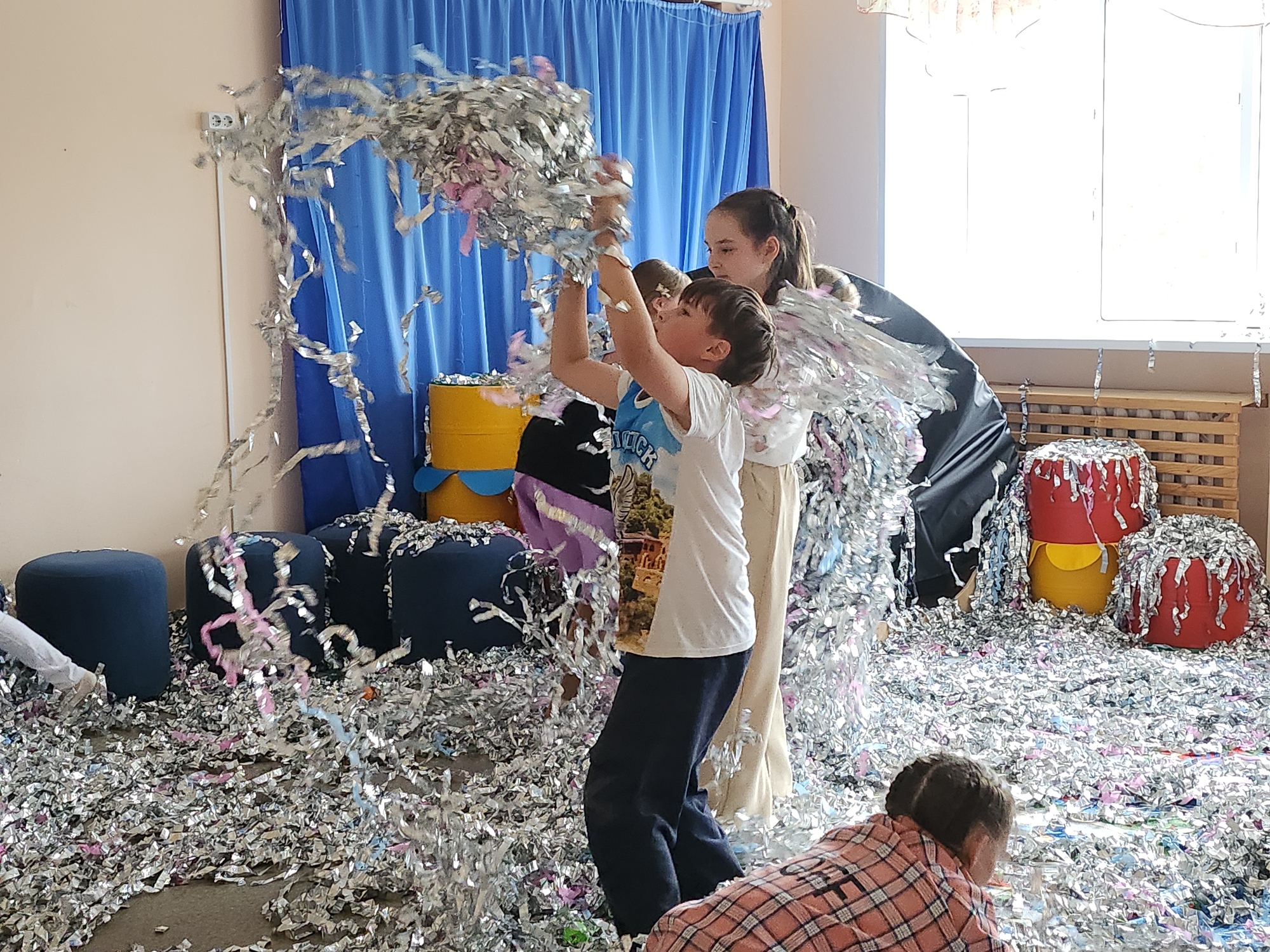 A group of children are throwing silver and pink confetti in an indoor space in what appears to be Krasnogorskoye, Russia. One boy, with dark hair, is in the center, arms raised and throwing confetti in the air. A girl with braided hair stands behind him, also throwing confetti. A third child is on the floor in the lower right corner, wearing a plaid shirt.
The background includes blue curtains, and several ottomans are covered in blue fabric, and some barrels are decorated with yellow and red paper, all covered in confetti. The floor is covered in silver and pink confetti. A window is visible on the right.