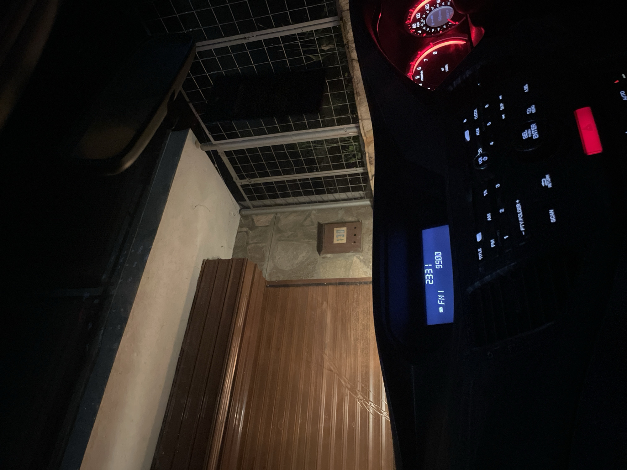 The image shows the view from inside a car looking towards what appears to be the exterior of a building. The car's dashboard and control panel are prominently featured, with the time displayed as 23:31 and the radio set to 99.00 FM. Outside the car, there is a wall and a door with vertical panels, under a partial metal grid. A sign reading 130 is on the wall next to the door. Based on the available details, it's not possible to provide the exact location in the city of Zlatograd, Bulgaria.