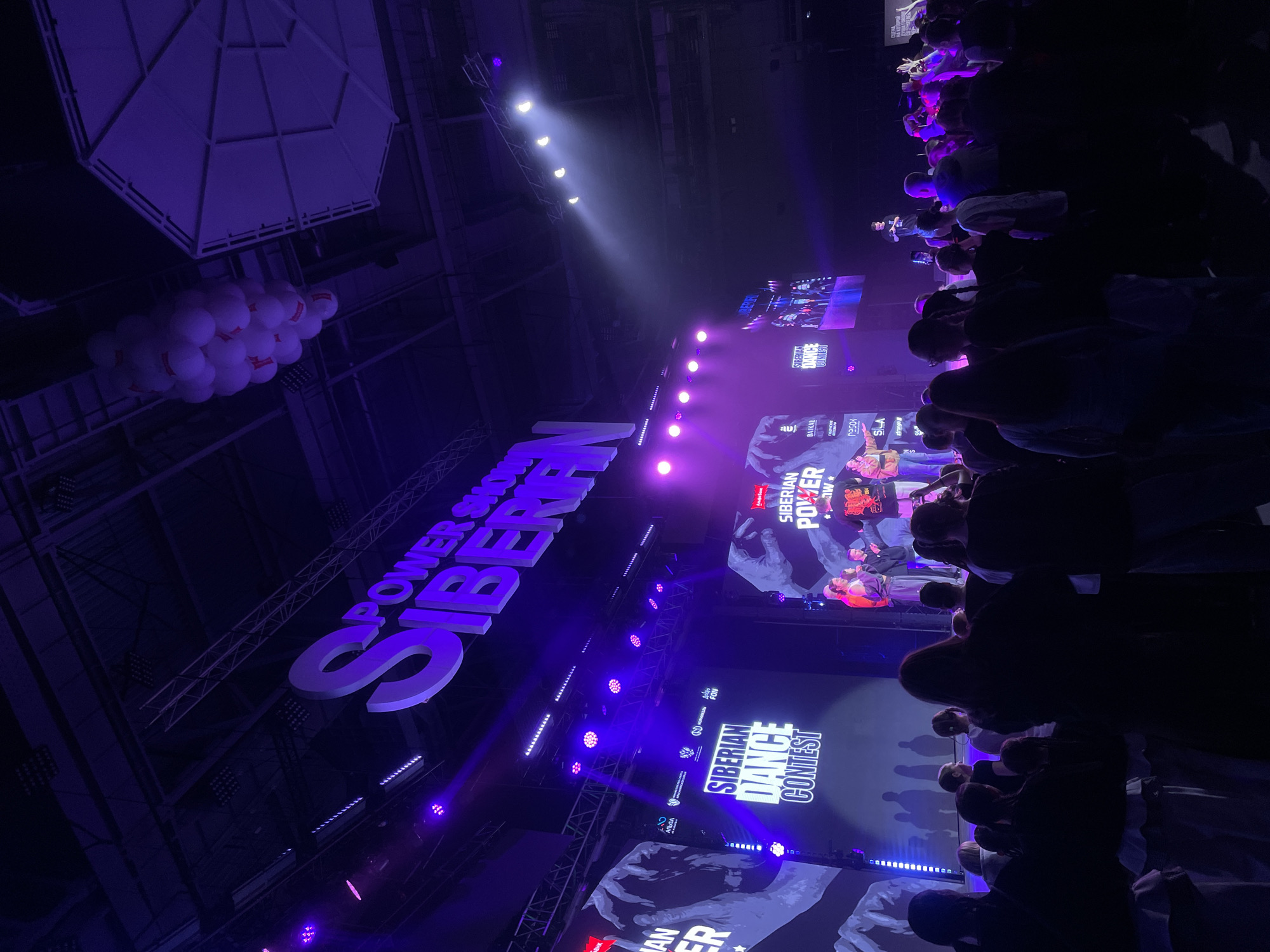 The image depicts an indoor event occurring at the Siberia International Exhibition Business Center (МВДЦ Сибирь) in Krasnoyarsk, Russia. The scene is dominated by a large stage illuminated with purple and blue stage lighting, featuring multiple large video screens. The screens display text such as "SIBERIAN DANCE CONTEST" and "SIBERIAN POWER SHOW," alongside graphic elements including stylized hands and mountain outlines, and various sponsor logos.
Above the stage, prominent three-dimensional white letters spell out "POWER SHOW SIBERIAN." Several individuals are visible on the stage, some actively presenting or interacting. In the foreground and midground, a substantial crowd of attendees, primarily seen from behind, faces the stage. The crowd consists of numerous individuals of varying ages and attire, largely silhouetted against the bright stage lights. Some attendees are actively engaged, with a few arms raised.
The venue features a high ceiling with exposed structural trusses and lighting rigs, from which numerous stage lights emit beams. A cluster of white balloons, each marked with a red symbol, hangs from the ceiling towards the upper right of the frame. The overall atmosphere suggests a dynamic, high-energy event.
