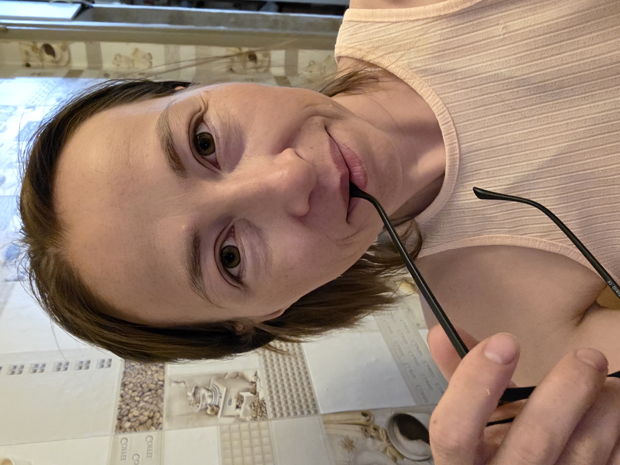 A woman with brown hair and brown eyes is holding a pair of black-rimmed glasses to her mouth, as if to bite them. She is wearing a sleeveless, ribbed, pale pink top. The background appears to be a kitchen or bathroom with tiled walls that have a pattern of coffee beans and cups. The lighting suggests an indoor setting.