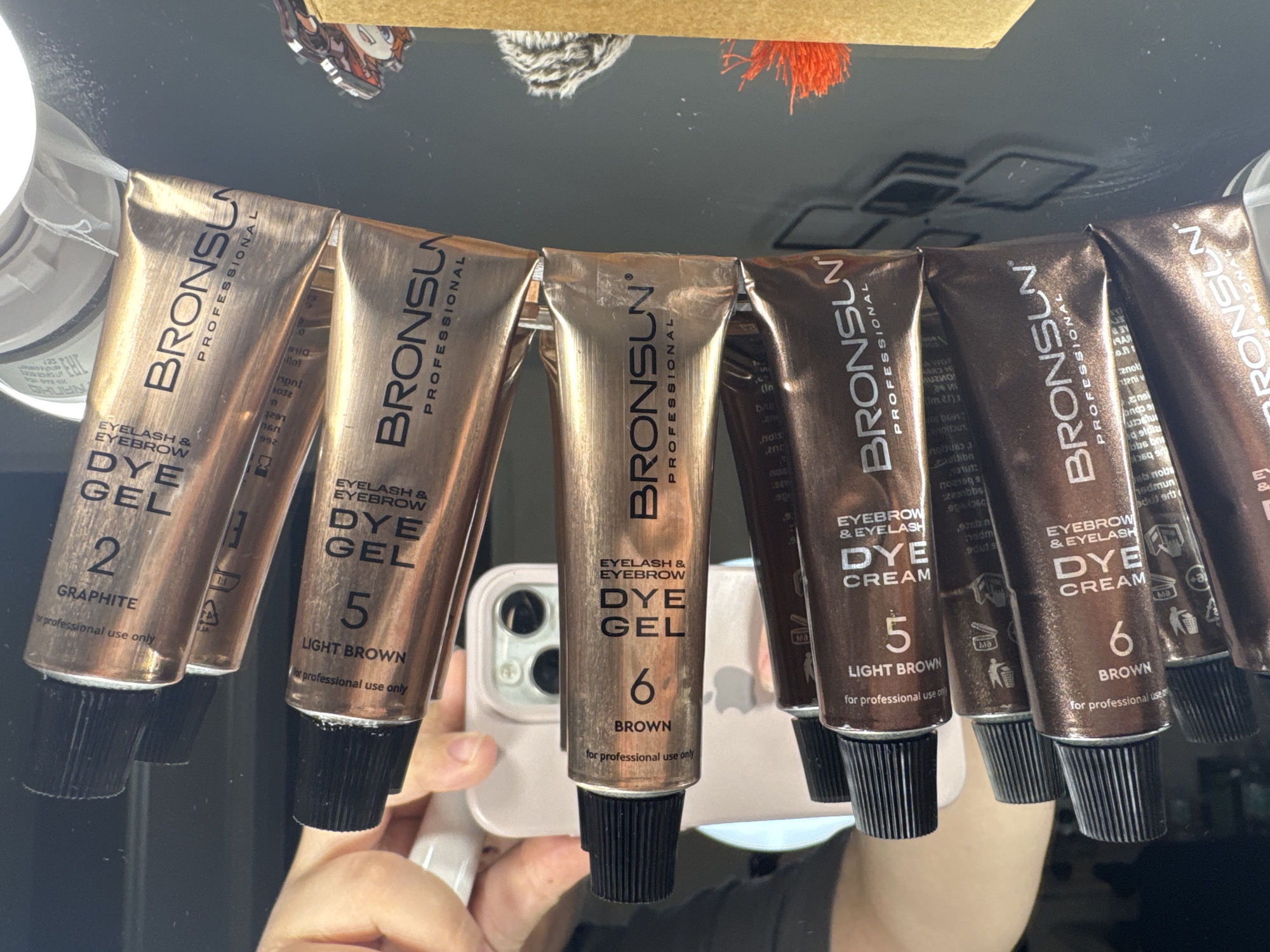 The image displays a collection of BronSun Eyelash & Eyebrow Dye Gel and Dye Cream tubes, arranged in a horizontal line. The tubes are primarily bronze and brown in color, with black caps. Visible text on the tubes includes "BRONSU" and "PROFESSIONAL," along with indications of shade numbers and names such as "2 GRAPHITE," "5 LIGHT BROWN," and "6 BROWN." Some tubes are labeled "DYE GEL" and others "DYE CREAM." The tubes are partially obscured by a person's hands and a mobile phone, which is pink and has a visible camera lens array. The background appears to be a reflective surface, possibly a table, with indistinct shapes and colors, including a blurry figure wearing a black shirt. The lighting suggests an indoor setting. There is no information or visual evidence within the image that can be used to determine the location in Saratov, Russia.