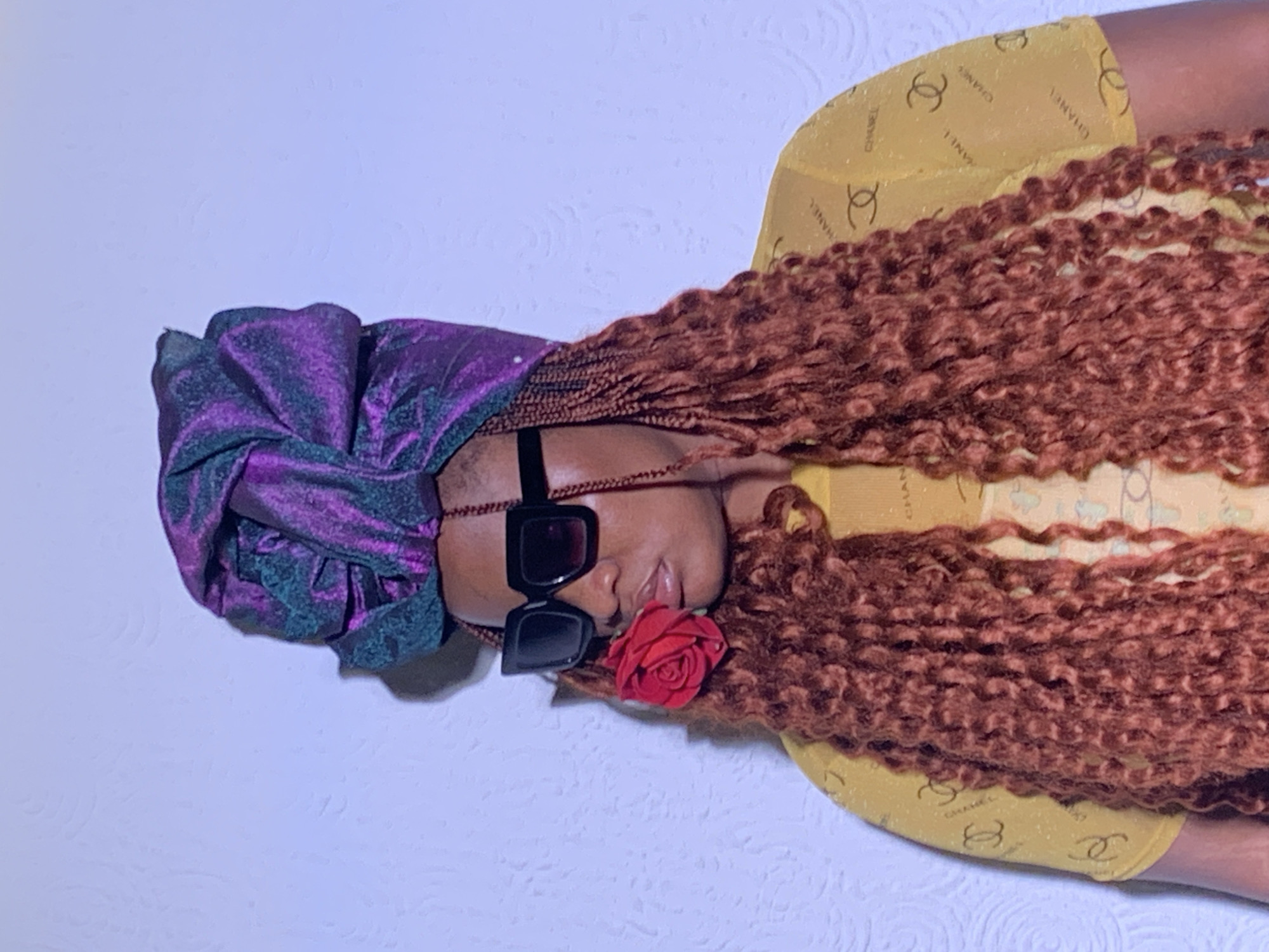 A person lies on a white textured surface. They wear a purple and green iridescent bonnet, large black square sunglasses, and a yellow shirt with Chanel logos. Brown textured hair spreads out. A red rose is positioned near their face. The location is Barakin Akawo, Nigeria.
