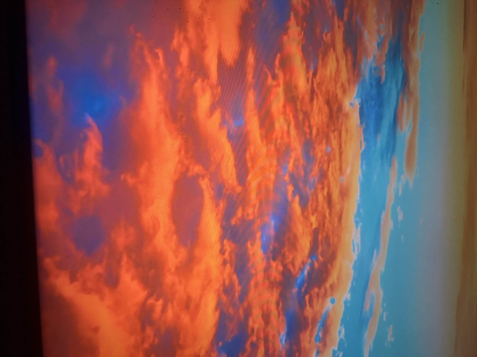 The image displays a dramatic sky filled with vibrant clouds, primarily in shades of orange and red, interspersed with patches of deep blue. The clouds are dense and textured, suggesting a sunset or sunrise. The lower portion of the image shows a clearer sky transitioning into a faint horizon, with hues of light blue and a hint of orange. There are no discernible buildings, people, or specific landmarks visible, making it impossible to determine a precise location within Herzliya, Israel. The scene is solely focused on the atmospheric conditions of the sky.