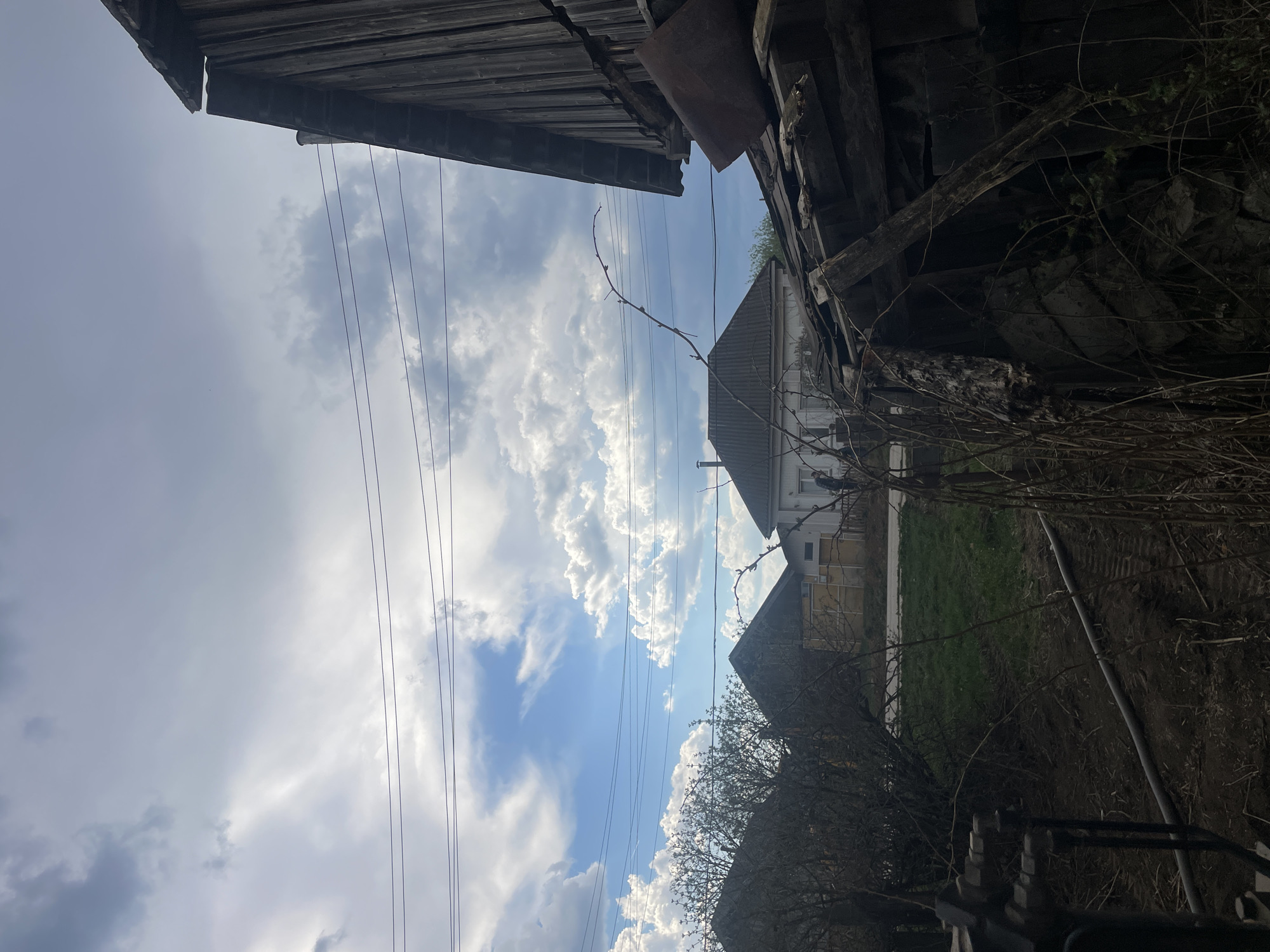 The image, taken from a low angle in Поедуги, Russia, shows a view upward towards a partly cloudy sky. Power lines stretch across the sky. Overhanging wooden structures, possibly parts of buildings or sheds, frame the top and right sides of the image. Two houses are visible in the middle ground, separated by a patch of green grass. In the foreground, bare tree branches and some obscured objects add depth to the scene. The overall impression is one of a rural or semi-rural environment with a mix of natural and man-made elements.