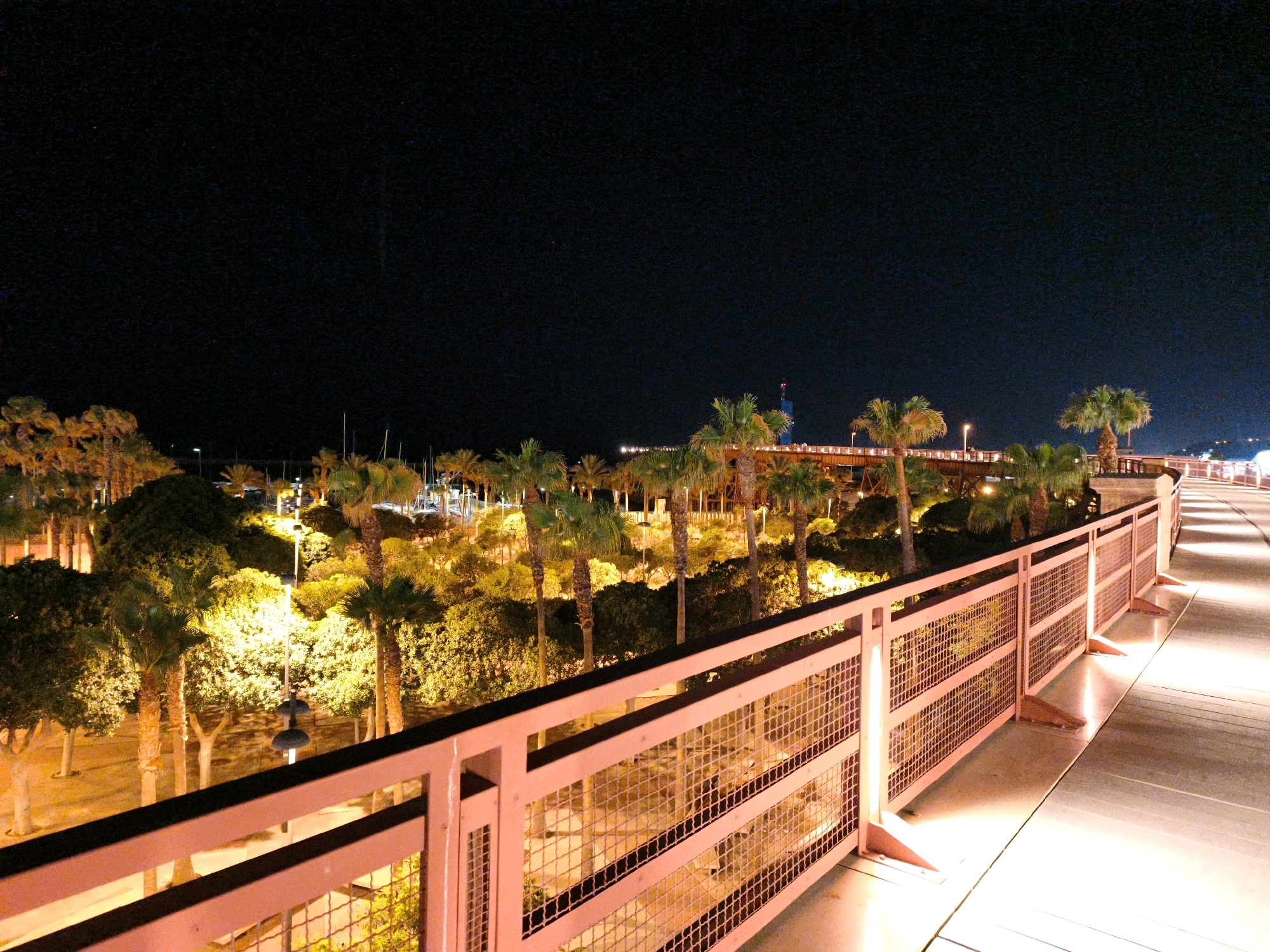 This image captures a nighttime view of a promenade or walkway in Almería, Spain. The foreground features a light-pink metal railing with mesh infill, separating the walkway from a densely vegetated area to the left. The walkway itself is illuminated by warm, yellow lights, casting long shadows and highlighting the textured surface. Beyond the railing, numerous palm trees and other foliage are visible, illuminated from below by artificial lights, creating a warm, inviting atmosphere. In the background, a marina with sailboats can be seen, along with distant buildings and structures, some of which are also lit. The sky above is a deep, dark black, indicating nighttime. The scene appears to be a well-lit public area, likely a tourist or recreational zone within the city.