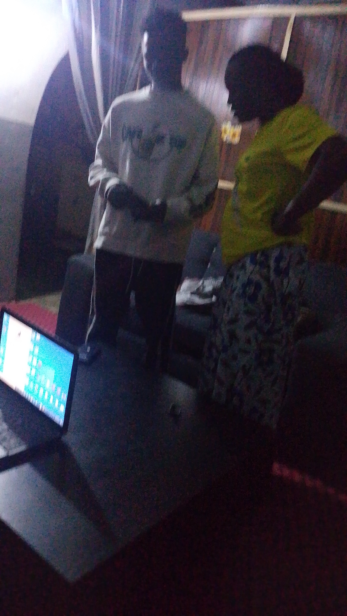Two individuals are standing in an indoor environment, possibly an office or living space, in the city of Unknown, Unknown. On the left, a person wearing a light-colored sweatshirt stands with their hands clasped in front. To the right, a second person is wearing a bright yellow t-shirt and a patterned skirt, with their back partially turned to the camera. In the foreground, a laptop is open on a dark surface, displaying a colorful screen. The background features vertical blinds and wooden paneling. The lighting is dim, creating shadows and a grainy appearance to the image.