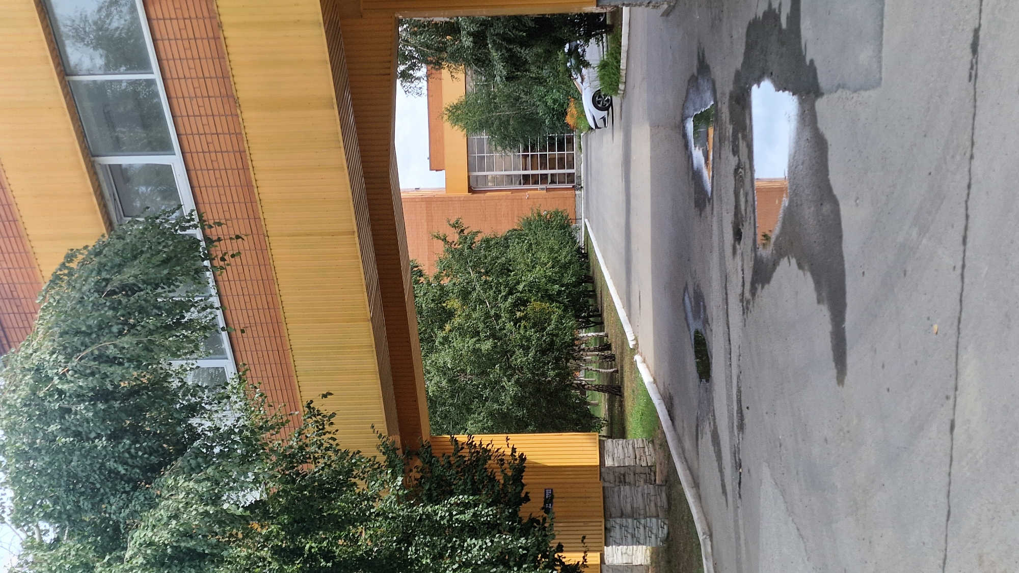 The image shows an exterior view of a building in Kostanay, Kazakhstan. The building is orange and yellow. A large overpass, also yellow, extends across the foreground. Trees line the pathway under the overpass. A white car is parked on the right side of the pathway. Puddles of water are on the asphalt in the foreground. The sky is overcast.