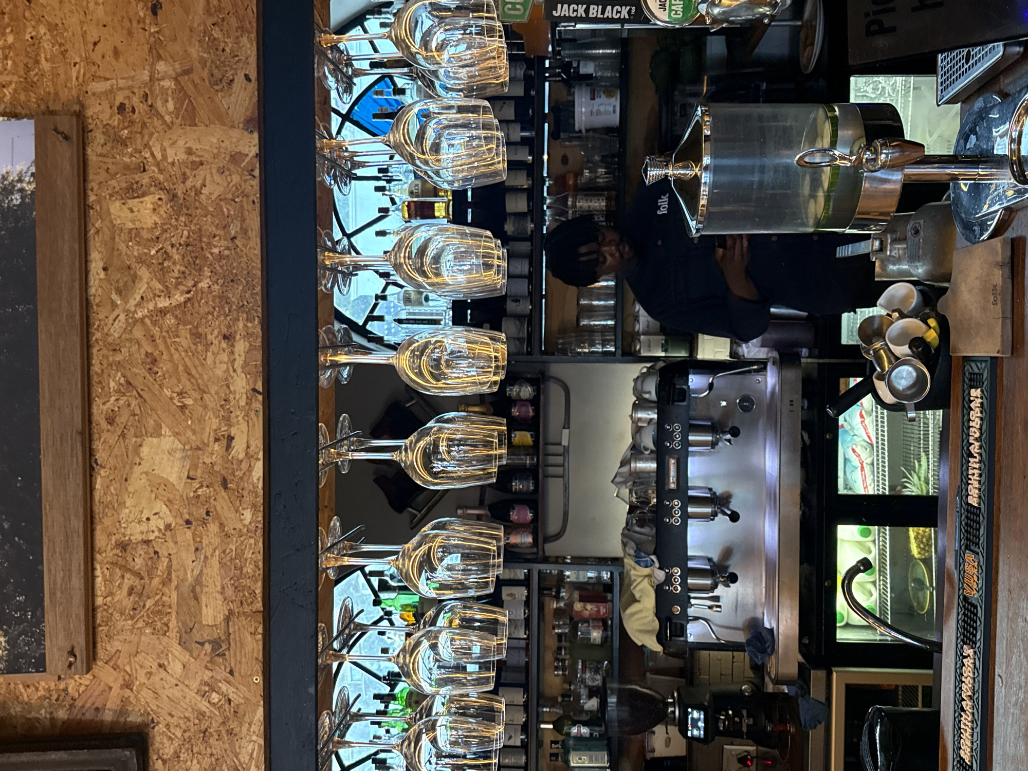 The image depicts a bar interior, likely in Cape Town, South Africa. The scene features a Black man wearing a dark uniform, potentially a bartender. He is standing behind the bar, seemingly interacting with equipment. A row of wine glasses hangs upside down above the counter. Behind the bar are bottles of liquor, coffee machines, and other bar equipment. The bar's aesthetic includes wooden paneling.