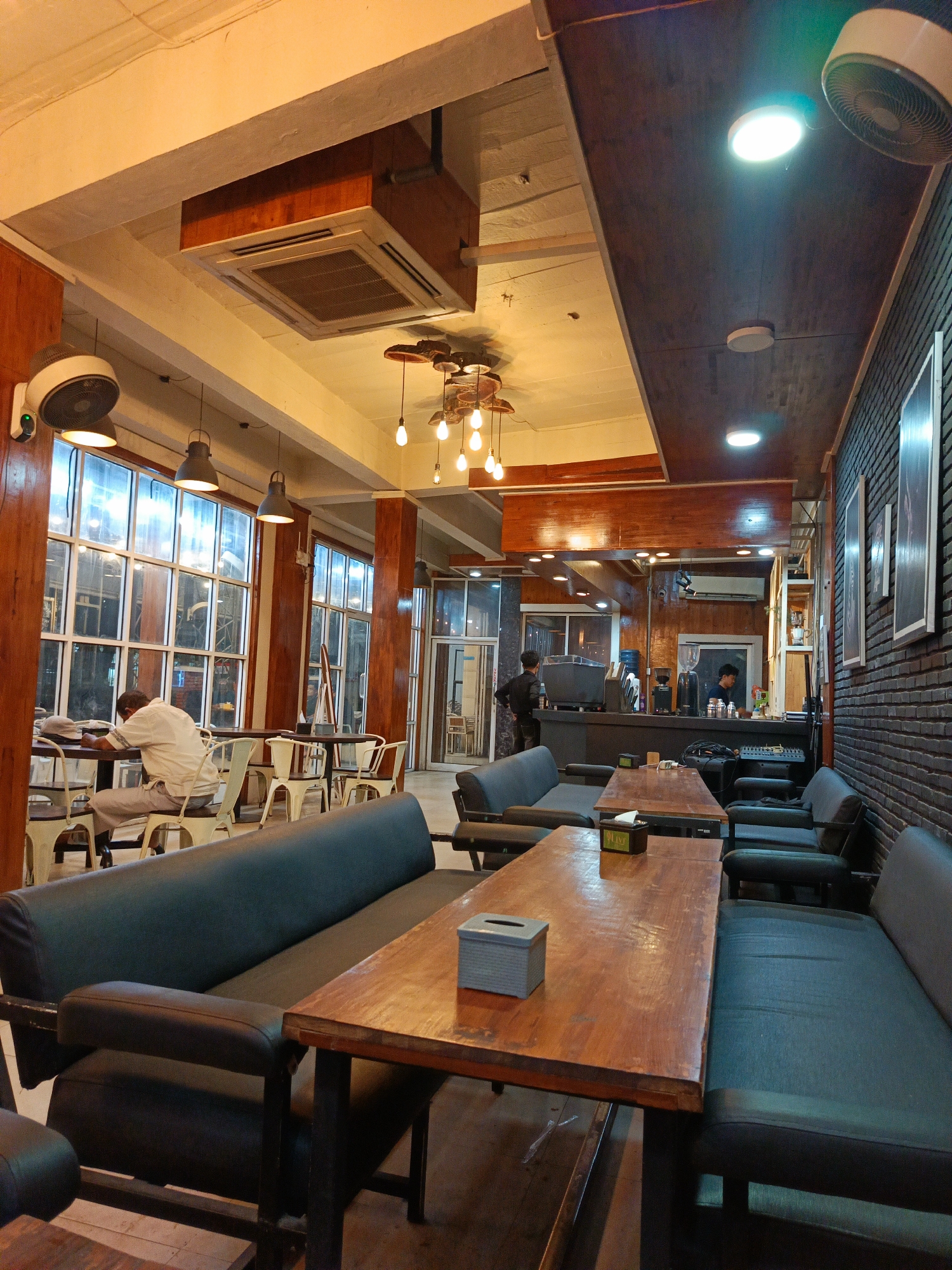 The image depicts the interior of a coffee shop, likely located in Lhokseumawe, Indonesia. The space is characterized by a mix of natural light filtering through large windows and artificial lighting from pendant lamps and ceiling fixtures. Wooden elements are prominent, seen in the ceiling beams, columns, and furniture, contrasting with the dark upholstery of the booth seating and the exposed brick wall.
In the foreground, several wooden tables are arranged with dark, upholstered booth seating on either side. A tissue box is visible on one of the tables. Further into the shop, a person is seated alone at a table by the window, with a hat placed on the table. This individual is facing away from the camera, appearing to be engaged in an activity, possibly reading or working.
Behind the seating area, a counter is set up with coffee-making equipment, including grinders and a coffee machine, and a water dispenser. Two individuals, presumably staff members, are visible behind the counter, one interacting with a customer and the other attending to tasks. The overall atmosphere appears to be relaxed and functional, suggesting it is a typical operational day at the coffee shop. The presence of an air conditioning unit mounted on the ceiling indicates efforts to maintain a comfortable environment for patrons.