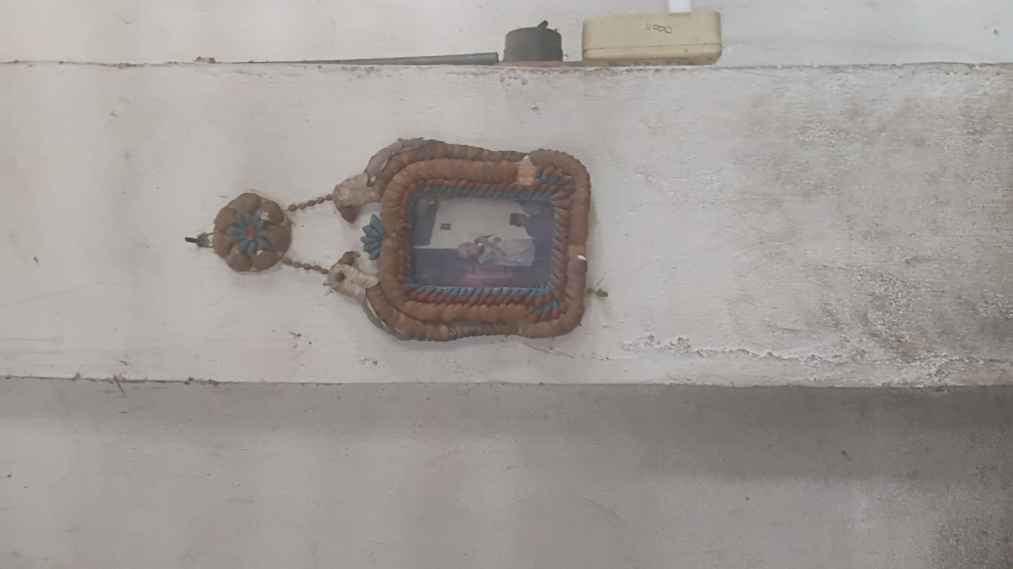 A framed picture hangs on a textured, off-white wall. The frame is rustic, made of braided material in earthy tones with blue and red accents, and features two decorative bird-like figures at the top. A floral-like ornament is attached above the frame. The picture inside depicts two individuals, one in the foreground with their face slightly obscured and holding an object, and another figure behind them who appears to be kneeling. The wall shows signs of age and wear, with some discoloration and rough patches. To the right of the frame, an electrical outlet and conduit are visible. The image composition suggests an indoor setting. The specific location within the city of Prabumulih, Indonesia, cannot be determined from the visual information provided in the image.