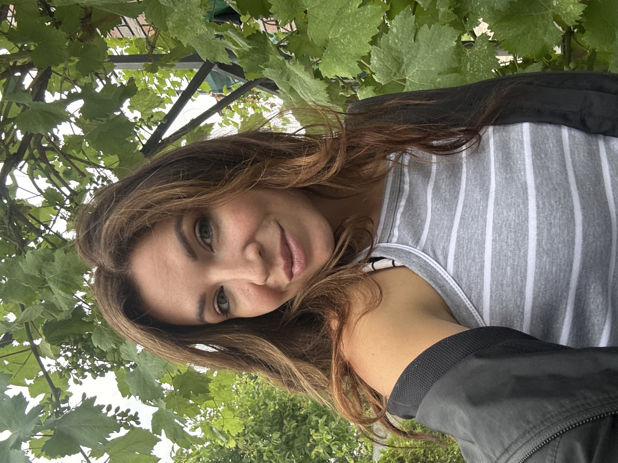 A young woman with long, wavy brown hair smiles into the camera, her head tilted to the left. She is wearing a gray and white striped tank top and a black jacket. The background is filled with lush green grape leaves and vines, suggesting an outdoor setting, possibly a vineyard or garden. The lighting is soft and diffused, indicating an overcast day. The location is identified as Lozuvatka, Ukraine.