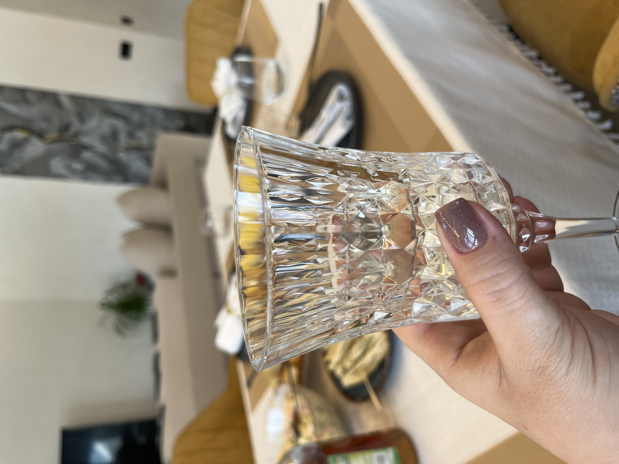 The image depicts an indoor dining setting in Новая Чернушка, Russia. In the foreground, a left hand, featuring shimmery mauve nail polish, holds a clear, faceted crystal stemmed goblet. The goblet is empty. The background reveals a dining table covered with a white tablecloth that has tassels, and beige placemats. Several place settings are visible, each with a dark plate, a white folded napkin, and cutlery. On the table, food items include a clear glass bowl of salad, a bottle containing red liquid with a green label, and a plate holding skewers of wrapped items. Yellowish-brown dining chairs are positioned around the table. The far background shows a light-colored sofa, a wall-mounted flat-screen television, a potted plant, and an abstract grey and gold wall art piece.