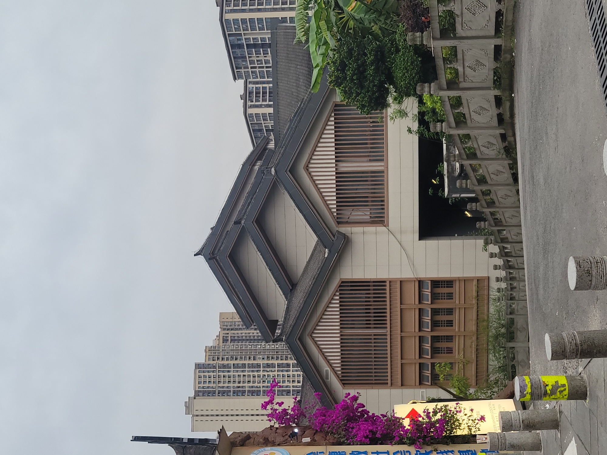 A view of a traditional-style building in Shapingba District, China, showcasing a distinctive diamond-shaped roof structure with grey tiles and light beige walls. Brown wooden window frames are visible. A high-rise apartment building rises behind the traditional building. A decorative grey stone railing runs along the sidewalk. In the foreground, a sign with purple bougainvillea flowers is visible. The sky is overcast. The scene is a street setting. Concrete bollards line the edge of the sidewalk.