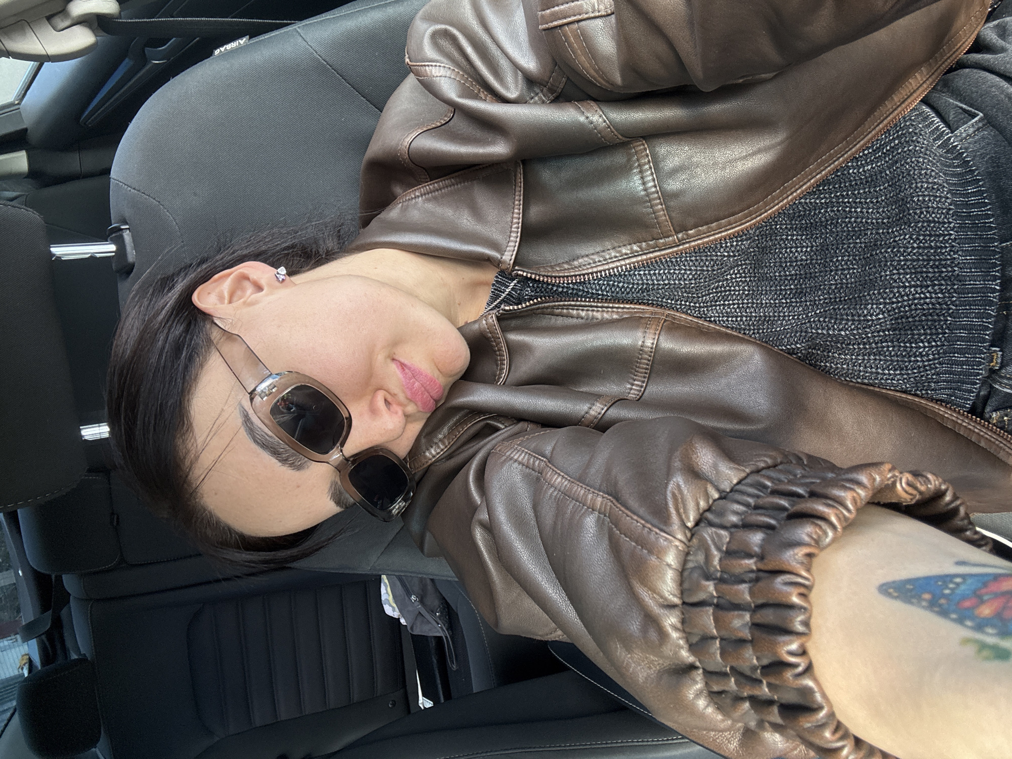 A person is sitting in the driver's seat of a car, wearing sunglasses and a brown leather jacket. The location is indicated by the headrest and safety belt of the car. Details include a tattoo on the person's arm and the urban environment visible through the car windows. The person is located in a car, likely in Tbilisi, Georgia.