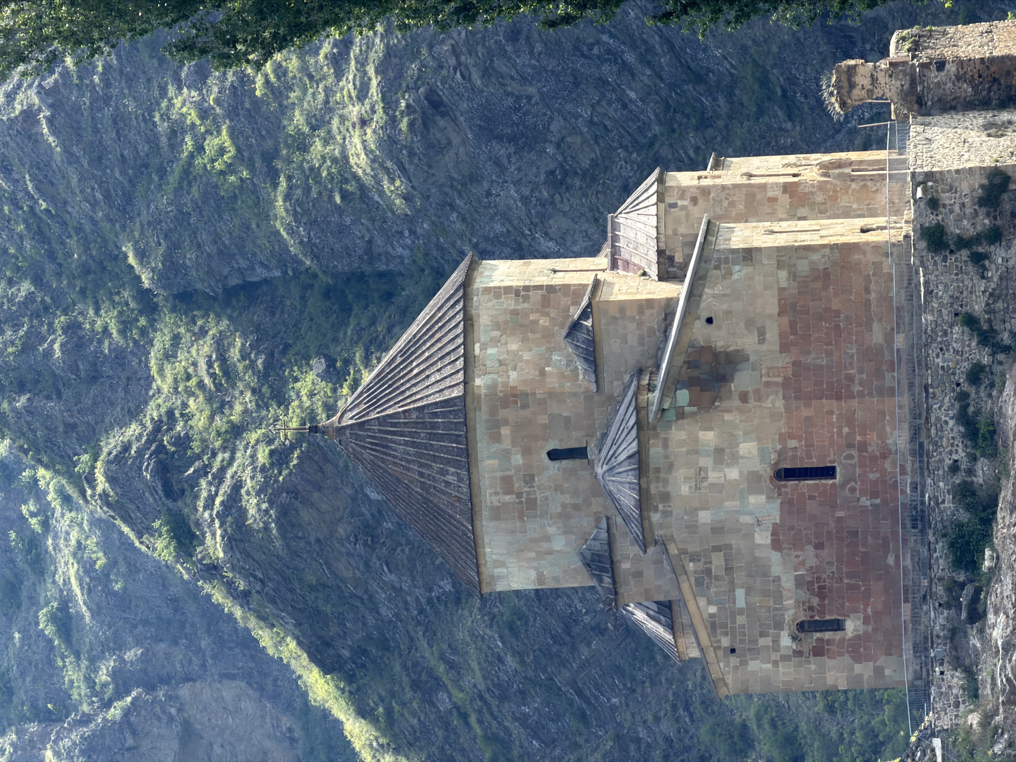 The image shows the Ateni Sioni Cathedral, a 7th-century Georgian Orthodox church located in the village of Ateni, near the city of Gori, Georgia. The cathedral is constructed from yellowish-brown sandstone and features a central dome supported by four columns. The exterior walls are adorned with carved stone reliefs, though these are not clearly visible in the image. The church is situated on a rocky hillside, with steep, vegetated slopes rising behind it. A tall, dark green tree is visible to the right of the cathedral. In the foreground, there is a stone wall and a walkway with a metal railing. The sky is a hazy blue. The scene is illuminated by sunlight, creating strong shadows and highlighting the texture of the stone and the landscape.