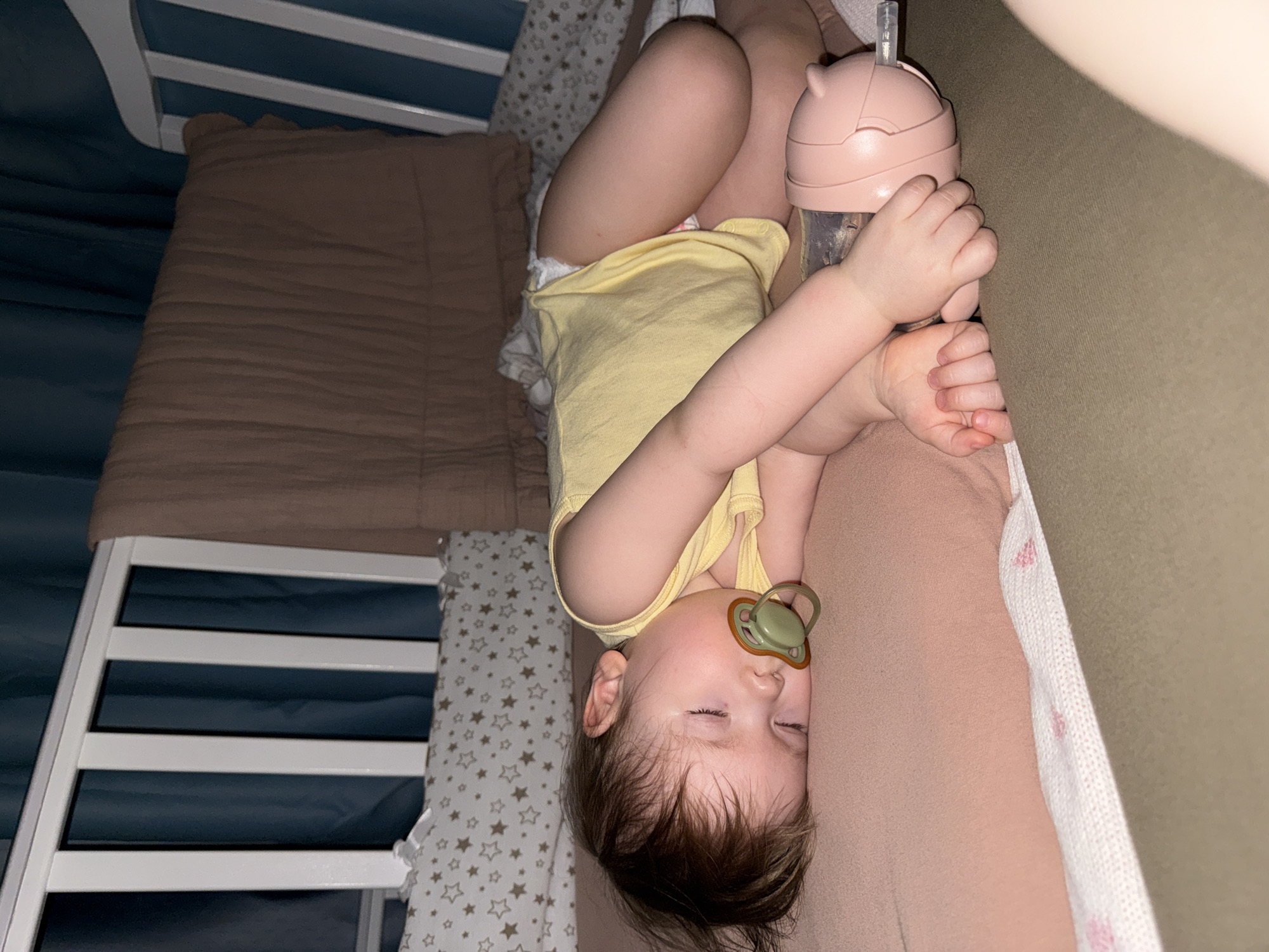 A baby is lying in a crib, holding a pink sippy cup. The baby has a pacifier in their mouth and is wearing a yellow shirt and a diaper. The crib has white bars, a brown pillow, and a bumper with stars. The location is not specified and not discernible from the image.