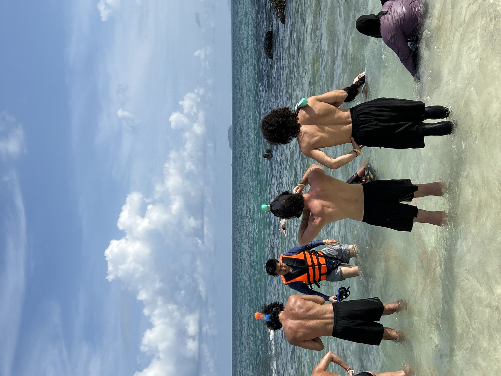In Phru Nai, Thailand, a group of individuals are standing in shallow, clear ocean water near a beach. The sky is blue with scattered white clouds. Several people are wearing snorkeling gear, including masks and snorkels. One person is wearing an orange life vest over a blue long-sleeved shirt and grey shorts. The water is calm and appears to be a tropical setting. The scene suggests preparation for or participation in water activities like snorkeling or swimming.