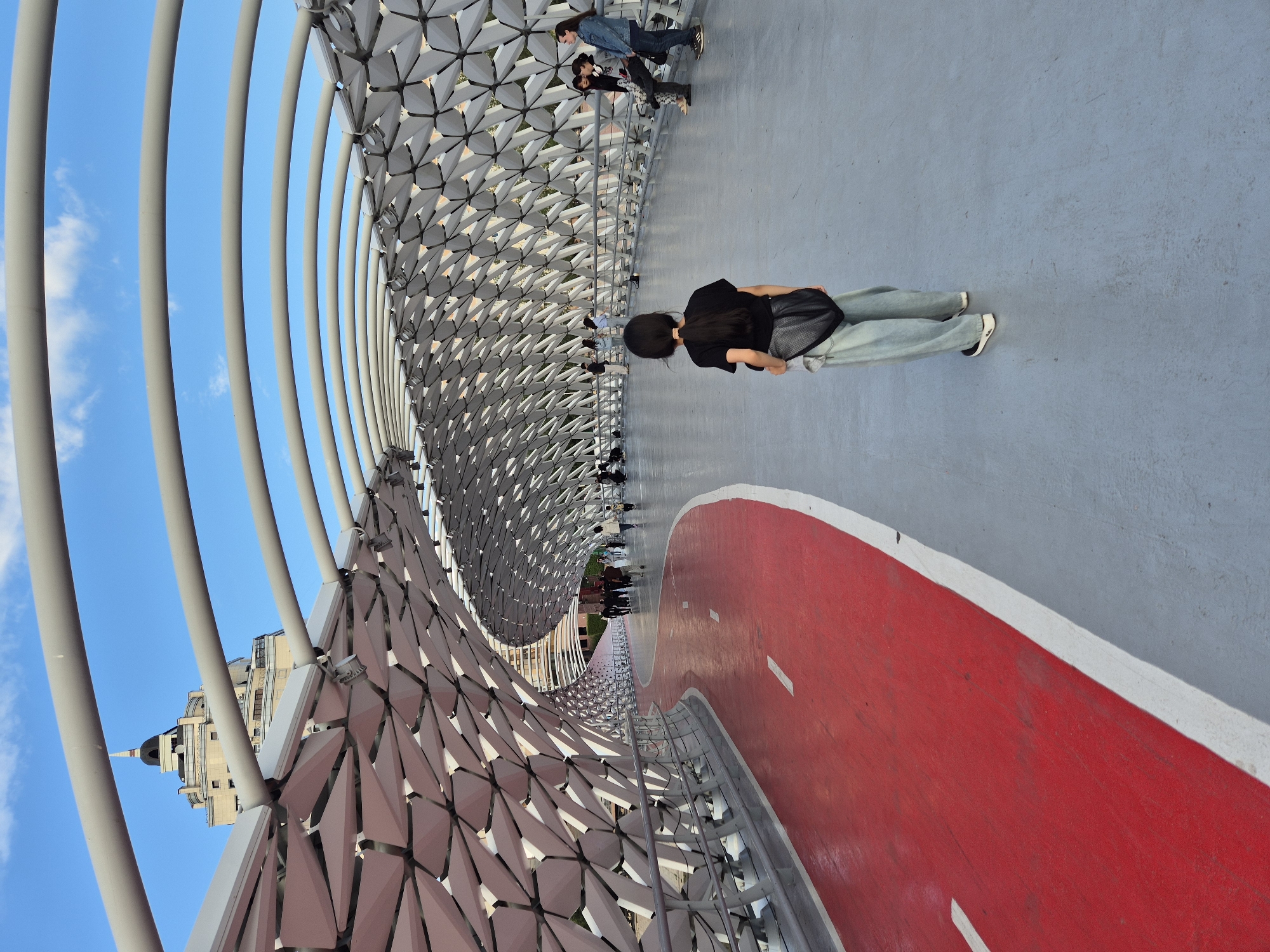 This is a full, eye-level, outdoor, vertical shot of the pedestrian bridge in Astana, Kazakhstan. The bridge is covered by a white, geometric, lattice-like structure. A red bike lane runs along the left side of the bridge, separated by a white line from the grey pedestrian walkway on the right. A young person with black hair is standing in the middle of the walkway, facing away from the camera, wearing a black t-shirt, a black mesh bag, and light blue jeans. Further down the walkway, several other people are walking and sitting. In the background, a tall, light-colored building with a dark dome is visible against a bright blue sky with scattered white clouds.