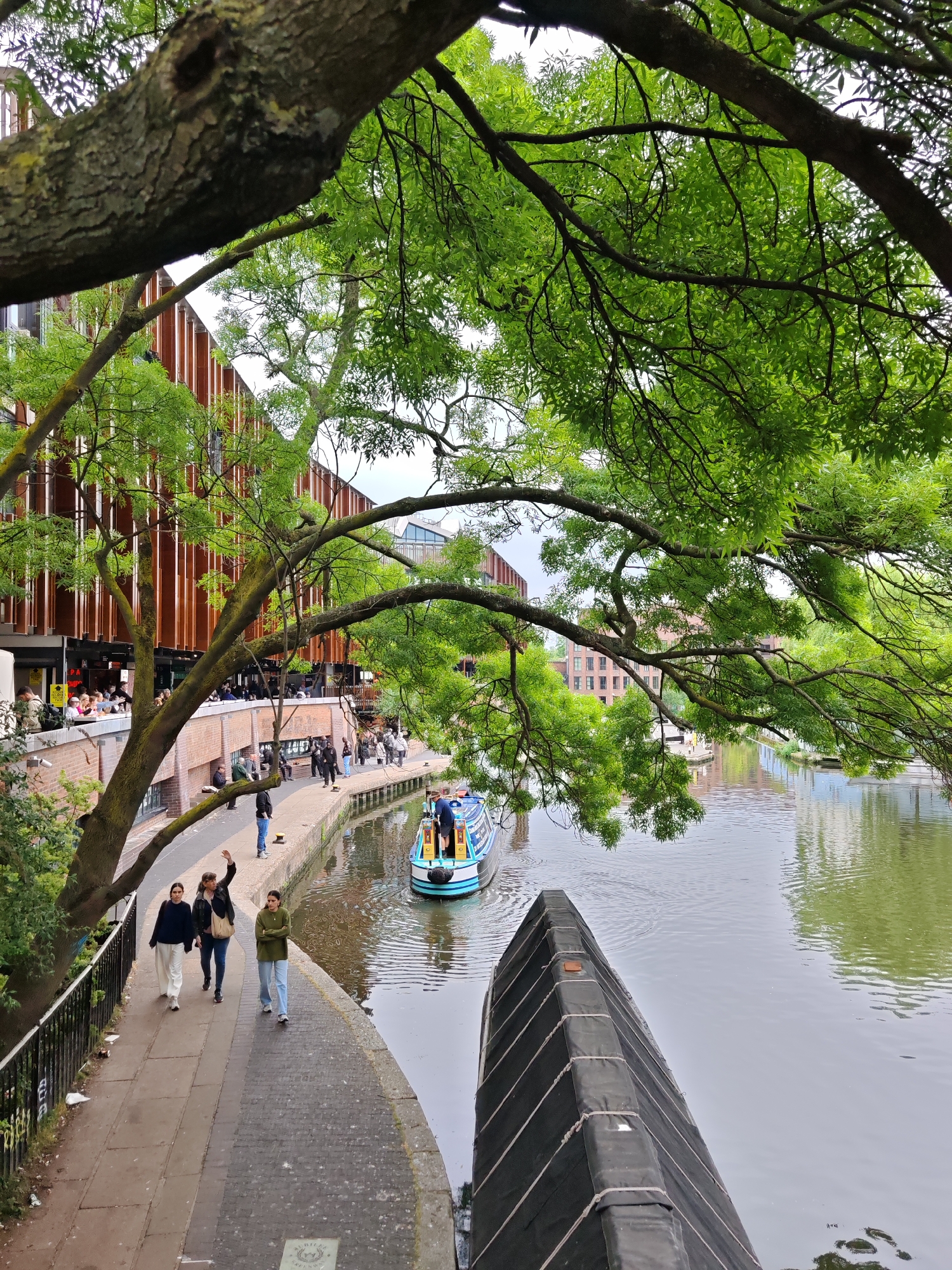 This image shows a canal in London, United Kingdom. The canal is flanked by a walkway on one side and various buildings, including ones with rust-colored cladding. On the walkway, three women are walking; one is waving. Further along the canal, a canal boat with people on board is visible. The scene is framed by tree branches with green foliage. A black object that is likely a canal boat cover or another floating fixture is in the foreground. Other pedestrians are present along the canal walkway. The weather appears to be overcast. The overall environment suggests a mix of urban and natural elements within the city.