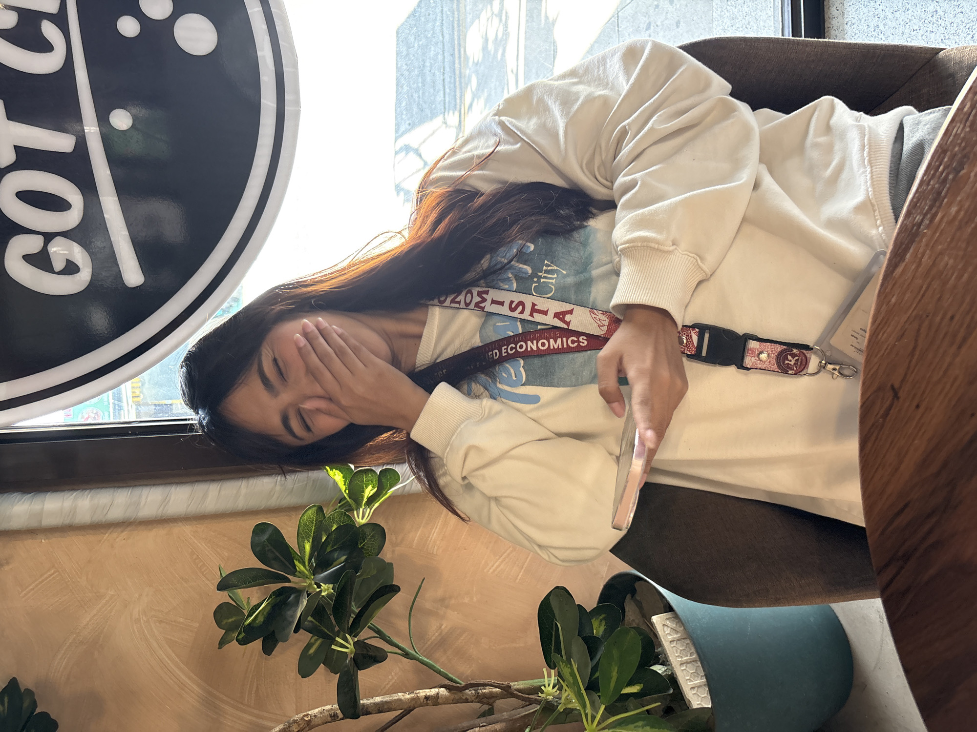 A young woman is seated in a dimly lit cafe, her laughter muffled by her hand as she clutches a smartphone. She wears a cream-colored sweatshirt with a blue graphic print and a lanyard with a university name. The cafe interior is warm-toned, with a textured wall and a green potted plant to her left. Outside the window, a blurred cityscape suggests a location in Davao City, Philippines. The overall impression is one of candid amusement and relaxed socialization.