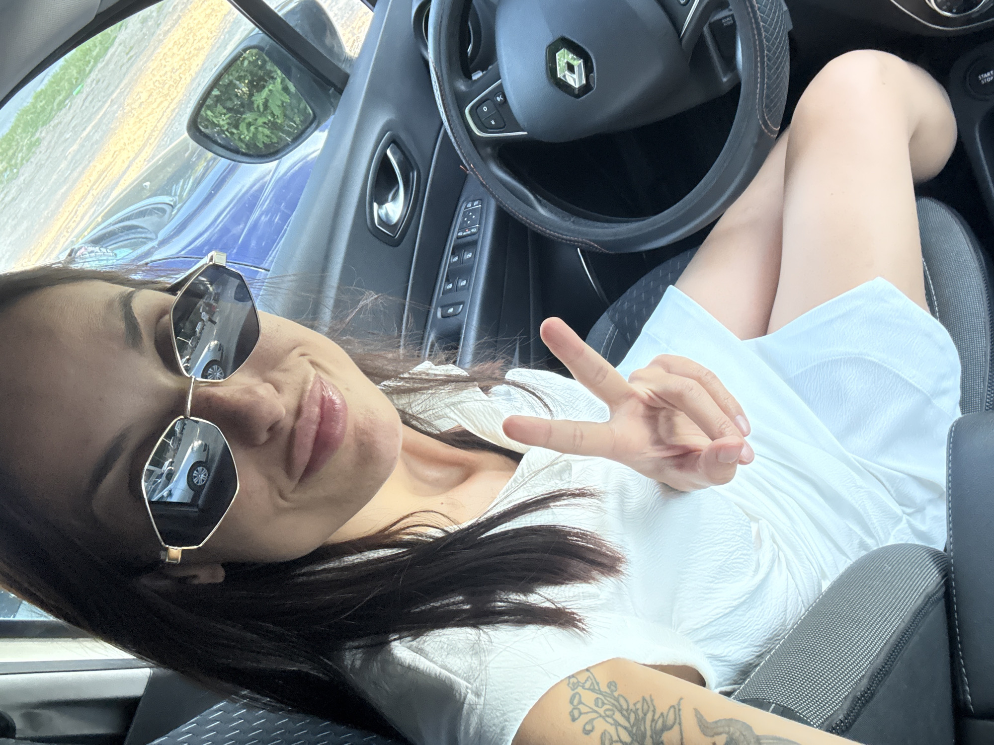 A woman wearing a white dress and sunglasses is sitting in the driver's seat of a car. She is holding up two fingers in a peace sign. The car's interior, including the steering wheel and door panel, is visible. The scene appears to be in a parking lot.