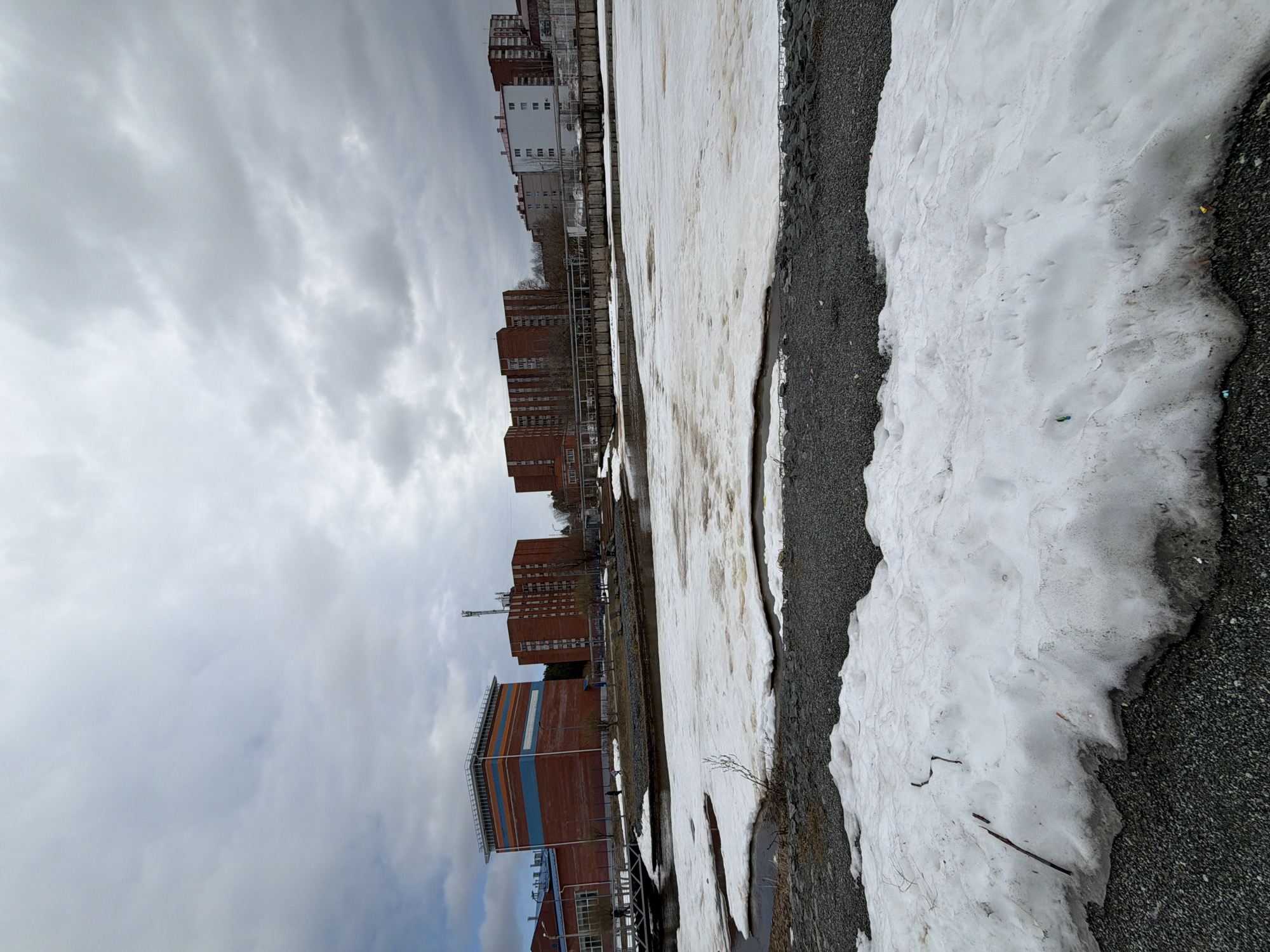 The image depicts a partially snow-covered landscape in Votkinsk, Russia, under a cloudy sky. Several brick buildings are visible in the background, including one with horizontal blue and orange stripes. A railway track runs parallel to the buildings. Patches of melting snow cover the ground near a gravel-covered area. The scene is mostly devoid of people or observable activities.