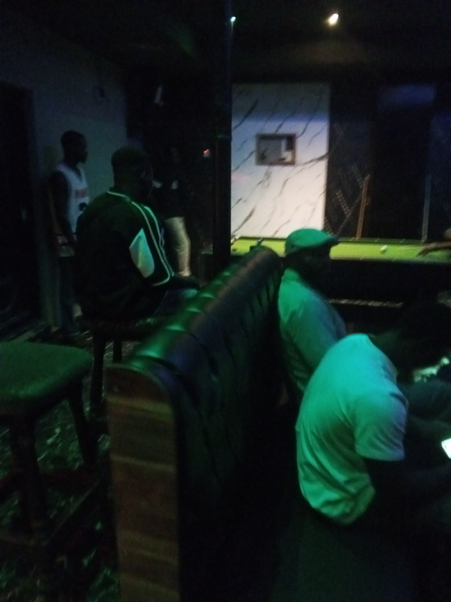 The image shows a dimly lit indoor scene with several people. In the foreground, a person wearing a bright green t-shirt is seated and facing away from the camera. To the left and slightly behind them, another person is seated, wearing a dark jacket with white accents. Further back and to the left, two other individuals are visible; one is wearing a white tank top and the other is mostly obscured. In the background, a pool table is set up with balls, and some decorative wall paneling is visible. The overall atmosphere appears to be a casual gathering in a recreational space.