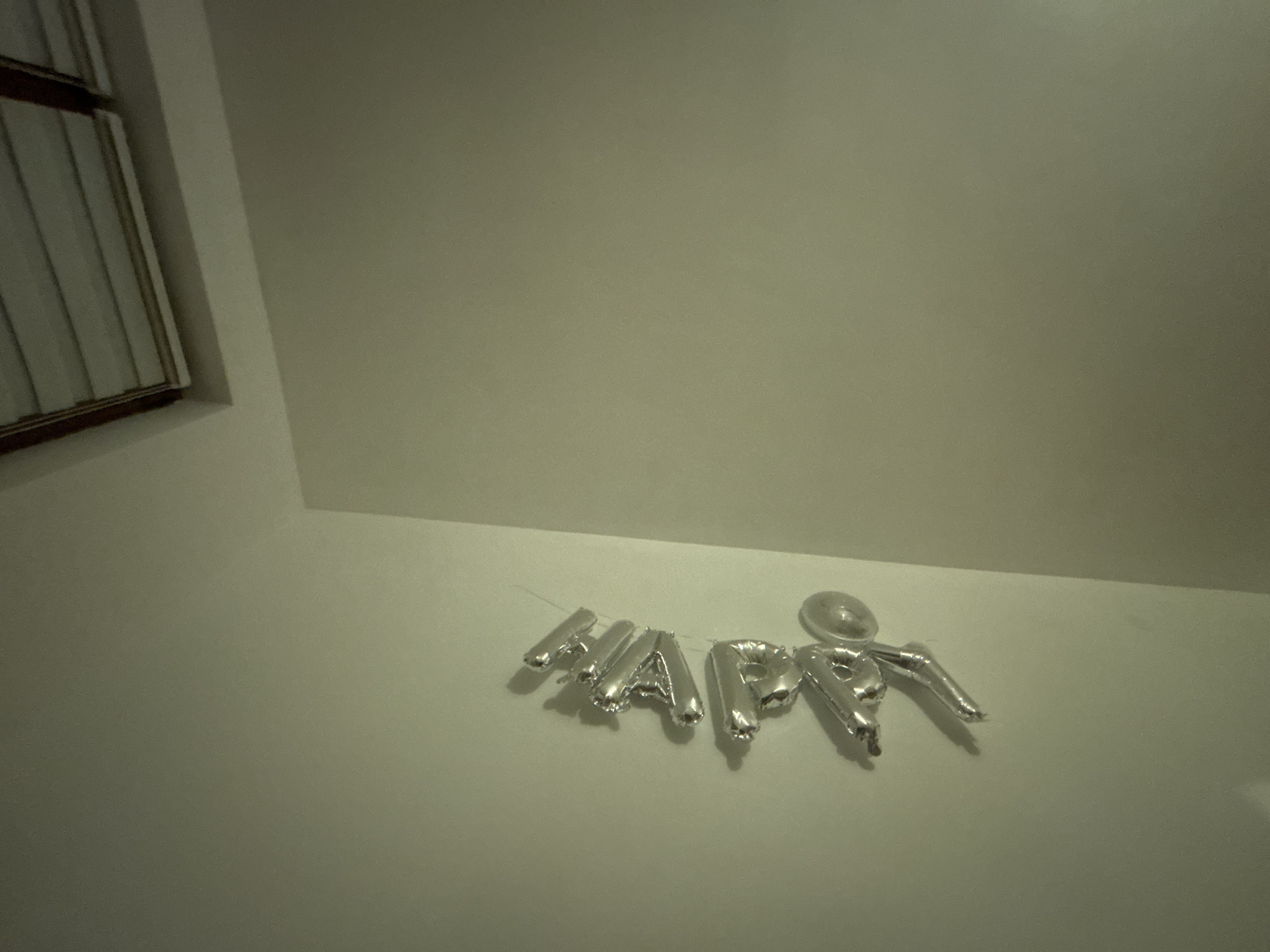 The image shows a silver "HAPPY" balloon decoration affixed to a light-colored wall. A window with closed blinds is visible in the upper left corner. The location is unspecified, but the image caption mentions Zlatograd, Bulgaria. There are no people or interactions visible. The environment is indoor.