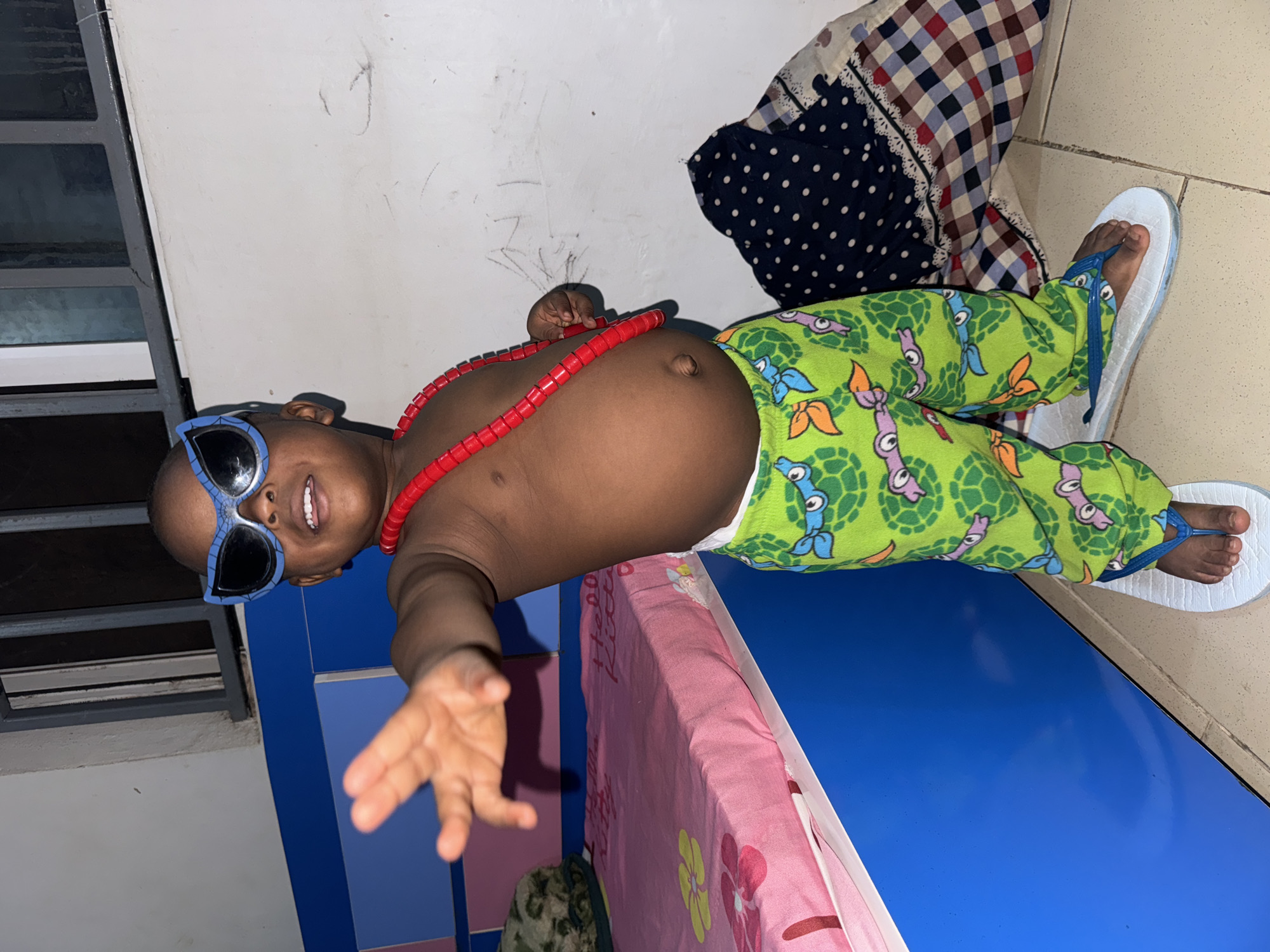 A baby wearing sunglasses, a red beaded necklace, and green patterned pajama pants is reclining indoors with white flip-flops on. The background includes a blue cabinet, a patterned blanket, a mattress with a floral pattern, a wall with scribbles, and a tiled floor. The city of Sabon Tasha, Nigeria, is not discernable from the image.