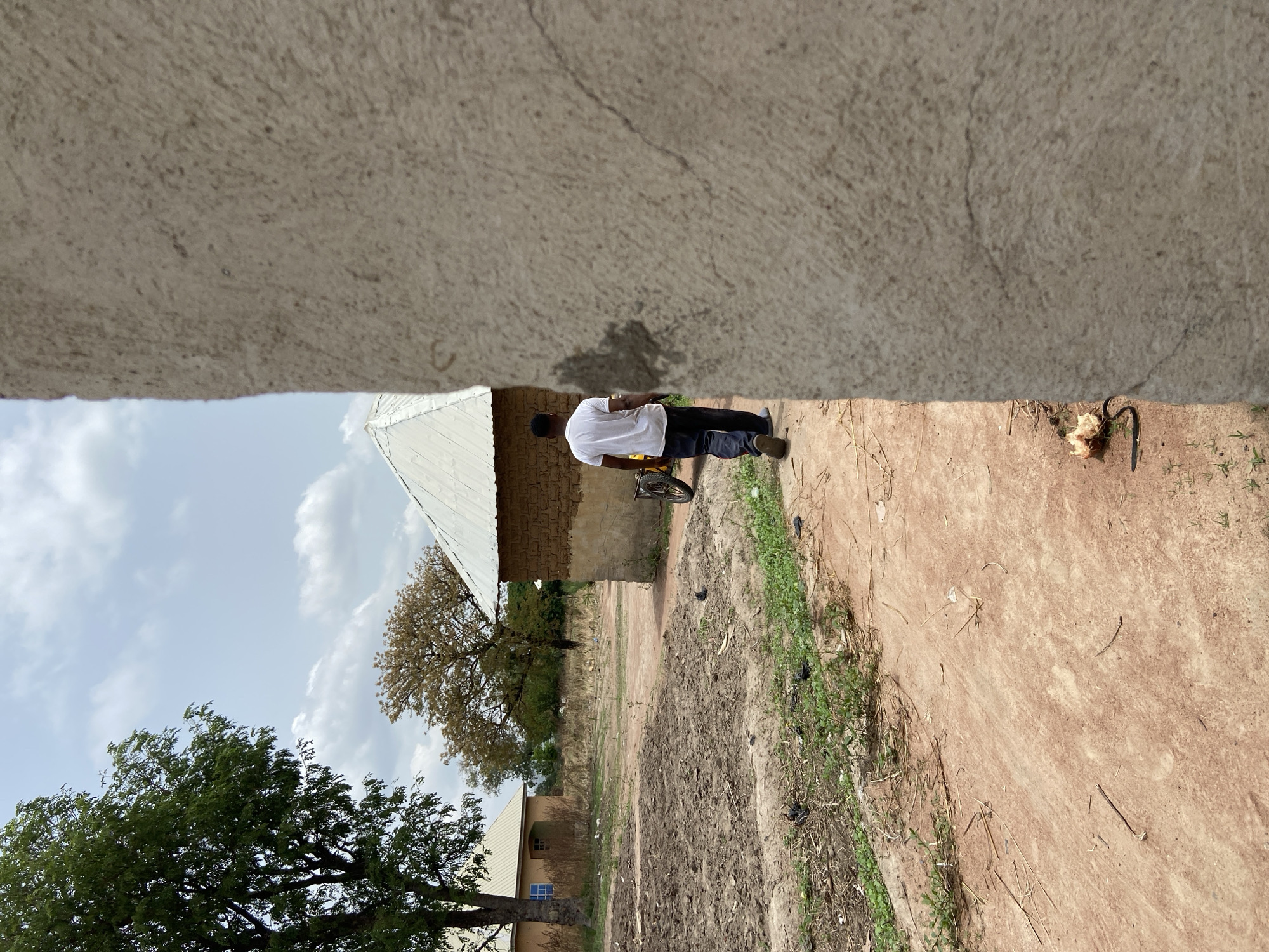 A daytime image depicts a semi-rural or peri-urban landscape in Mararaba, Nigeria, captured from a slightly elevated perspective looking across a dirt path. A large, out-of-focus, light-colored concrete or plaster surface dominates the top of the frame, suggesting the camera is positioned under an overhang.
In the mid-ground, a man is visible from behind, dressed in a white t-shirt, dark trousers, and light-colored shoes. He stands or walks beside a bicycle, part of its yellow wheel and frame visible. Behind him, several single-story buildings feature white corrugated metal roofs. The closest building has light brown, textured walls, possibly constructed from mud-brick or rendered. To the left, a large, lush green tree stands prominently, while another, possibly drier tree is further in the background. The ground is a reddish-brown dirt path, interspersed with patches of green vegetation and dry straw, showing signs of foot traffic and tire marks.
The sky is a light blue with scattered white clouds, indicating clear daytime weather conditions. Near the bottom right corner, a dark, bent metal object and a light-colored piece of debris are present on the ground. The scene conveys a sense of everyday life in a modest, unpaved environment. No discernible text is visible in the image.