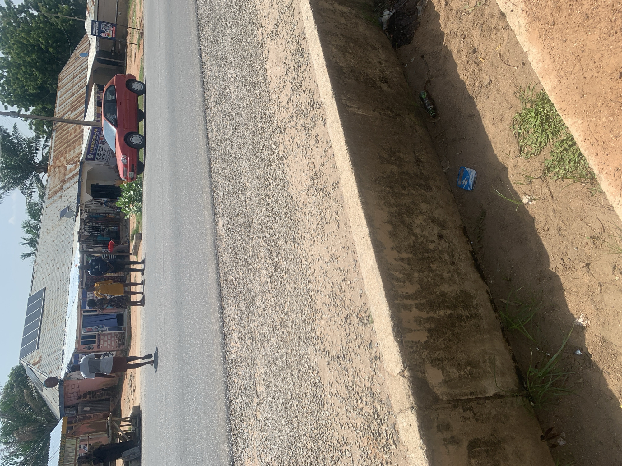The image depicts a street scene in Jalingo, Nigeria. A man in a grey and white shirt and a brown skirt is walking away from the camera across a paved road. Several people, appearing to be three women and one man, are standing on the side of the road near a row of shops with corrugated metal roofs. One of the shops has "HEBRON-M GLOBAL CONCEPT" visible on a sign. A red hatchback car is parked on the side of the road. The foreground of the image shows a concrete curb and a dirt ditch with sparse vegetation and some litter, including a blue and white packet and a discarded bottle. The environment appears to be a commercial area with modest buildings.