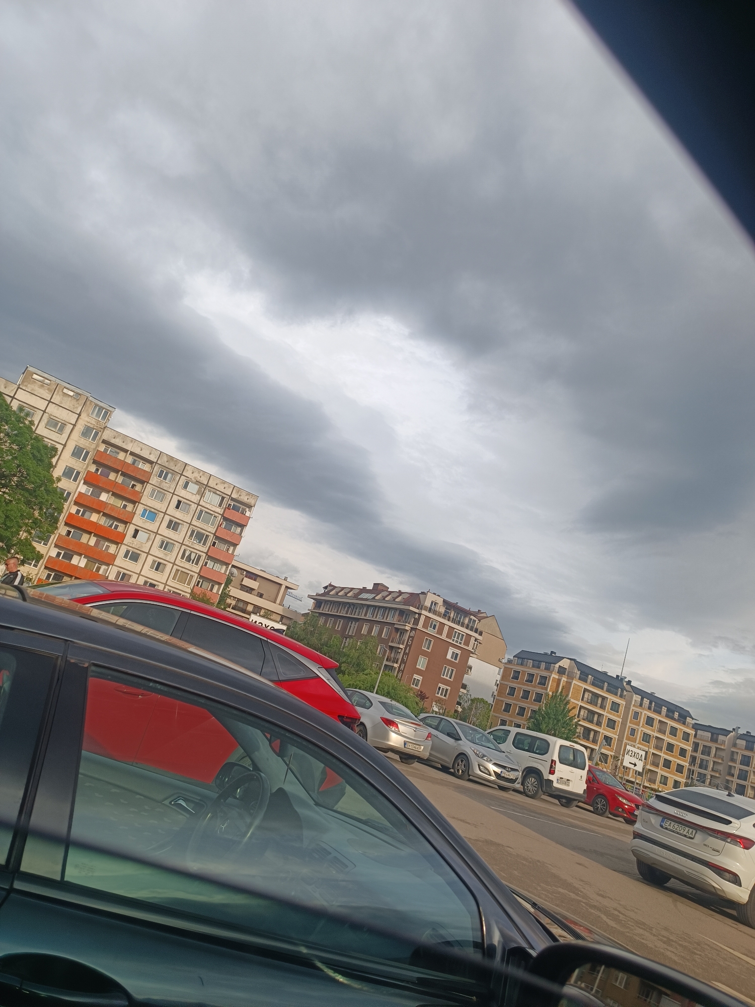 The image depicts a parking lot in Sofia, Bulgaria, with multiple cars parked in rows. In the foreground, the interior of a dark-colored car is visible, with the steering wheel and dashboard reflected in the window. Through the window, other cars are seen, including a red hatchback, a silver sedan, a white van, and a white SUV with license plate EA 6209 AA. Behind the cars, several multi-story residential buildings are visible. The sky is overcast with dark, heavy clouds. The overall environment suggests an urban setting during the daytime.