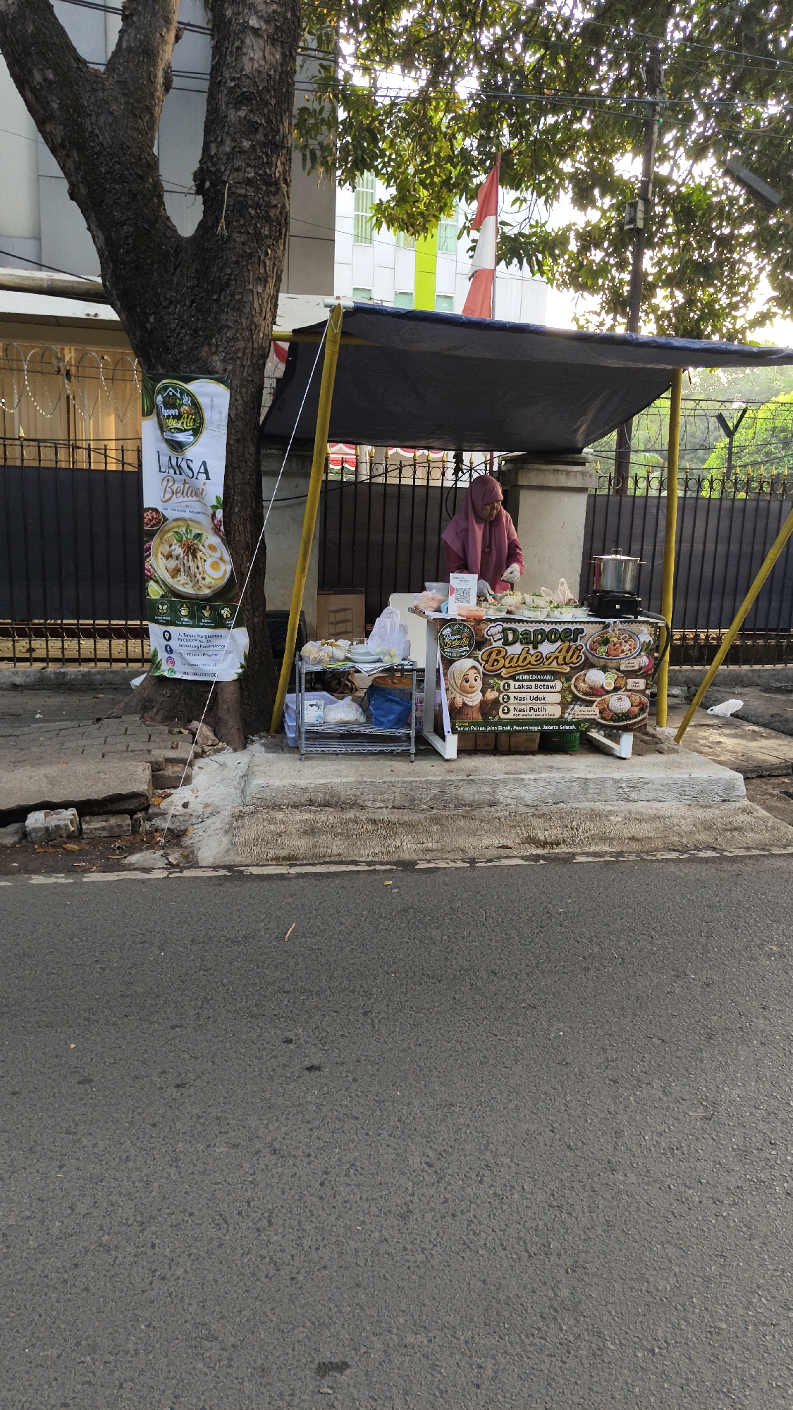 A street vendor is preparing food at a stall located on a sidewalk in South Jakarta, Indonesia. The stall, covered by a dark tarp supported by yellow poles, displays a banner for "DAPOER BABE ALI" with images of food items such as laksa, nasi udak, and nasi putih. The vendor, wearing a pink hijab and top, is behind the counter, working with ingredients. Next to the stall, a metal shelf holds packaged goods. A banner advertising "LAKSA Betawi" is attached to a tree on the left side of the frame. The background shows a black metal fence and a building. The street in the foreground is paved and appears to be a road.