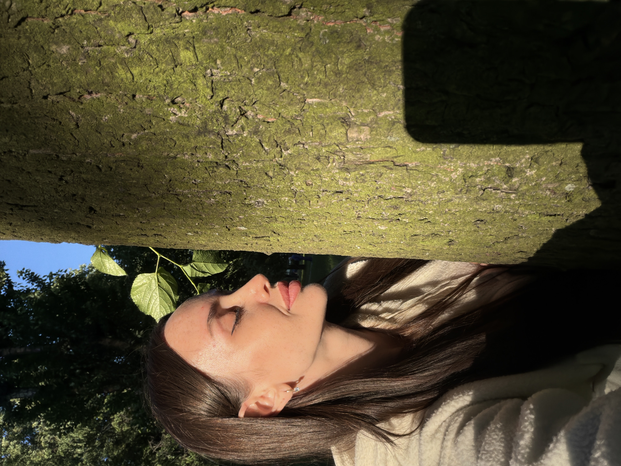 The image is a medium close-up shot of a woman with long, dark hair and fair skin. She is positioned to the left of a large tree trunk, with her head turned to the right, looking towards the tree with her eyes closed and a slight smile on her lips. She is wearing a cream-colored fuzzy jacket. The tree trunk is rough with patches of green moss. Sunlight illuminates the woman's face and hair, casting a warm glow. The background shows a blurred view of trees and a clear blue sky, suggesting an outdoor setting in a park or natural area. The overall lighting indicates it is daytime, likely late afternoon or early morning due to the angle of the sun. The specific location in Saint Petersburg, Russia cannot be determined from the image.