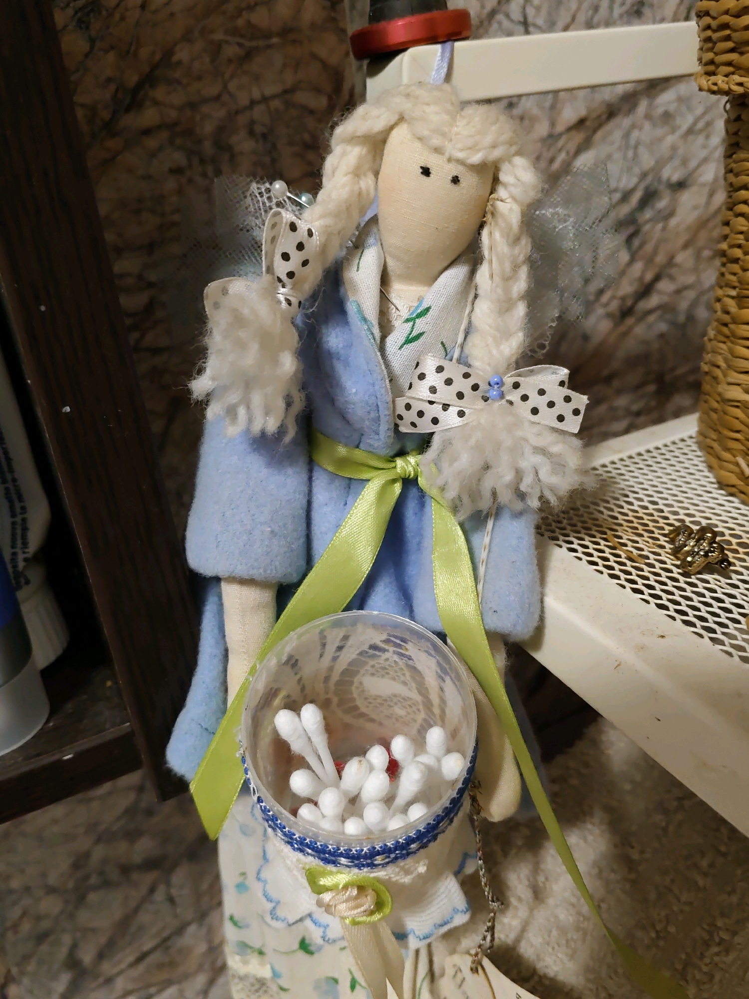 A handmade doll, adorned with a light blue coat and a green ribbon, stands upright. The doll has blonde yarn hair and two black button eyes. It holds a clear plastic cup filled with cotton swabs. The doll is positioned on a shelf in what appears to be a workshop or storage area. A woven basket is visible to the right of the doll. The background features a textured wall and shelves containing various items, including tubes of what appear to be toiletries or art supplies.