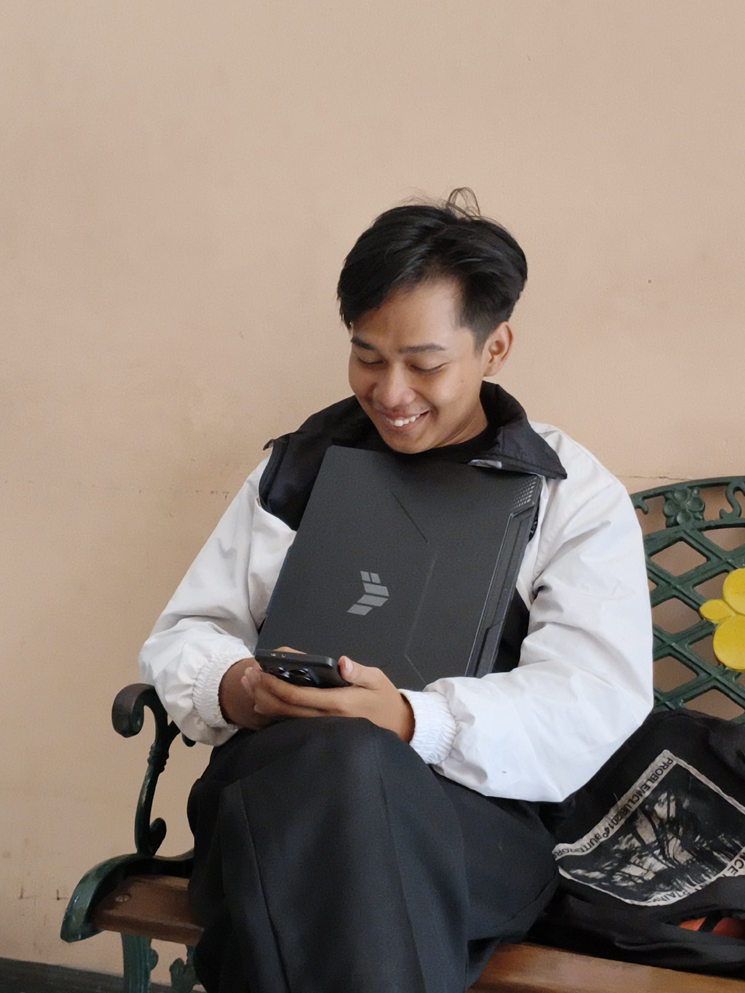 A young man is seated on a green metal bench with yellow accents. He is holding a black laptop with a distinctive logo and a smartphone. He wears a white jacket over a black shirt and black pants. His hair is dark and neatly styled. The background is a plain beige wall. A black bag rests on the bench next to him. He is smiling and looking at his phone. The scene appears to be located in Cianjur, Indonesia.