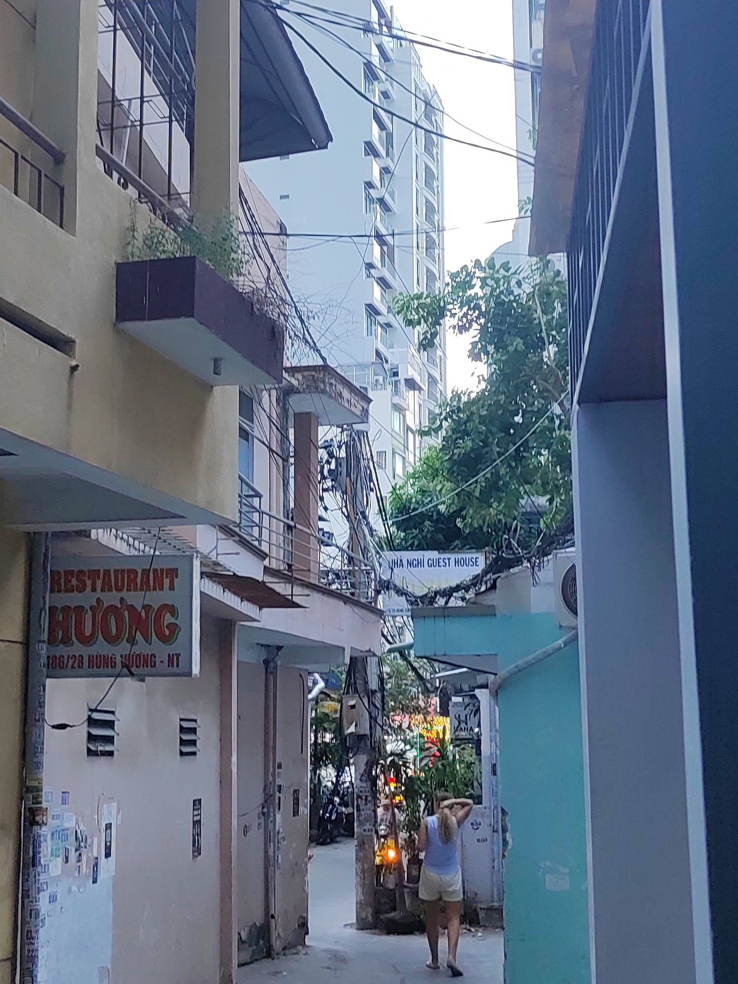 This is a medium shot of a narrow street in Nha Trang Ward, Vietnam. A woman is walking away from the camera down the street, wearing a light blue tank top and yellow shorts. To the left, a sign reads "RESTAURANT HUONG" and below it "186/28 HUNG VUONG - NT". Further down the street, a sign reads "NHÀ NGHỈ GUEST HOUSE". Buildings line both sides of the street, with wires and cables strung between them. A tall, modern building is visible in the background. The street is paved, and there are some plants and lights visible.