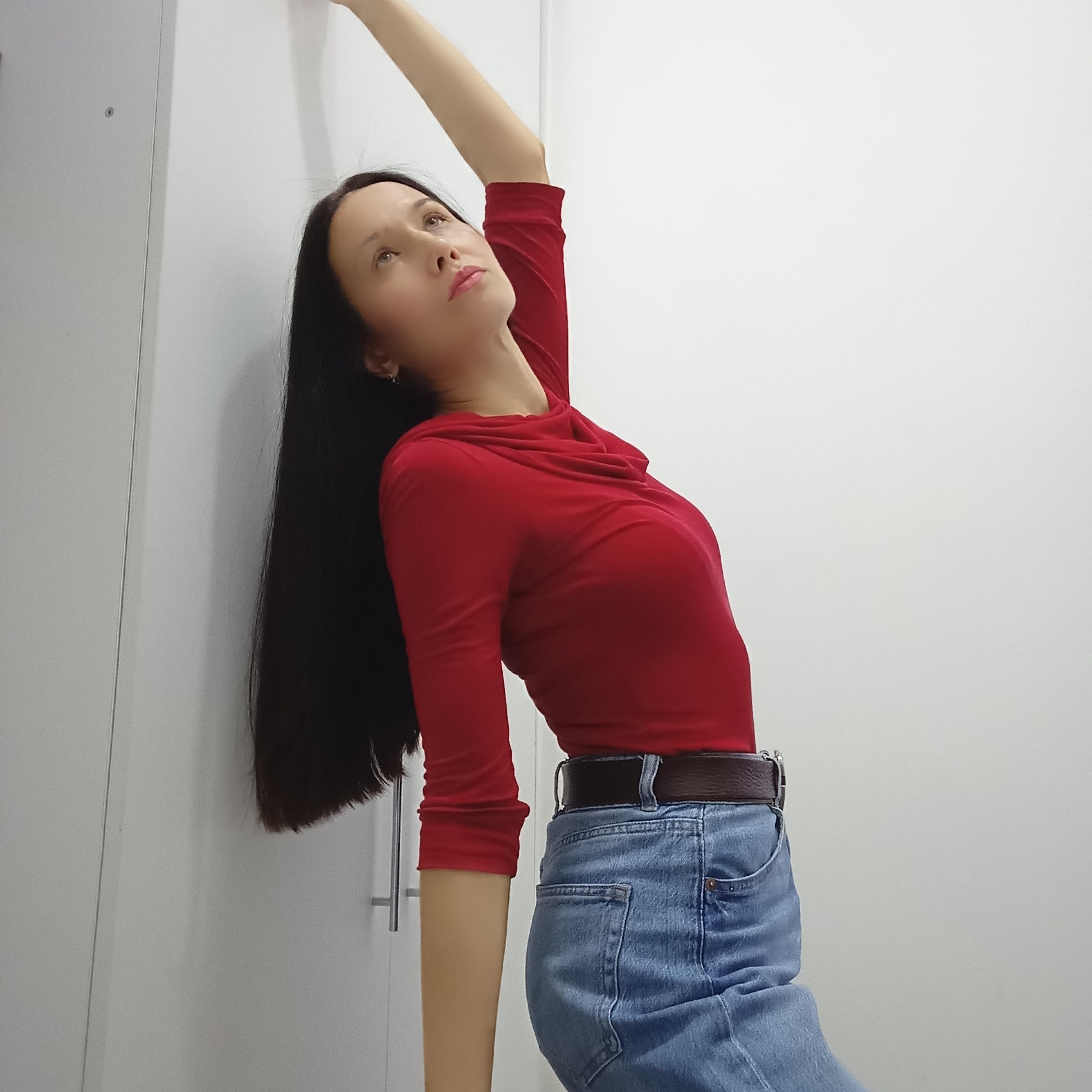 A young woman with long, dark hair is depicted indoors, leaning against a plain white wall or wardrobe. She is wearing a bright red long-sleeved top with a cowl neckline and light blue denim jeans secured by a dark brown belt. Her body is arched backward, with her head tilted upwards and her gaze directed towards the ceiling, conveying a serene or thoughtful expression. Her right arm is extended upwards, partially out of the frame, while her left arm hangs alongside her body. The background consists of minimalist white surfaces, featuring vertical seams and a silver handle on the left side, indicating a contemporary indoor setting. The lighting is bright and even, suggesting daytime. No visible text is present in the image. The location context for the image is Izhevsk, Russia.