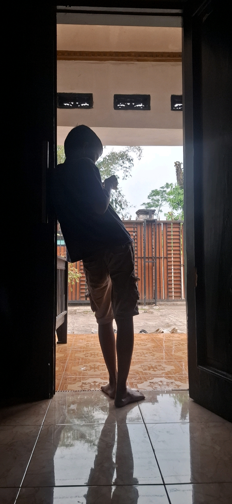 A person, silhouetted against the bright outdoors, leans against a dark doorway. They are wearing a dark t-shirt and light-colored cargo shorts. Their feet are bare. The person appears to be holding a small object, possibly a mobile phone, in their hands. The doorway opens onto a tiled patio. Beyond the patio, there is a wooden fence and lush green trees, suggesting a residential area in Cibinong Hilir, Indonesia. The sky is overcast. The polished tiles reflect the person's shadow and the surrounding environment. The overall impression is one of a quiet, casual moment captured in an outdoor setting.