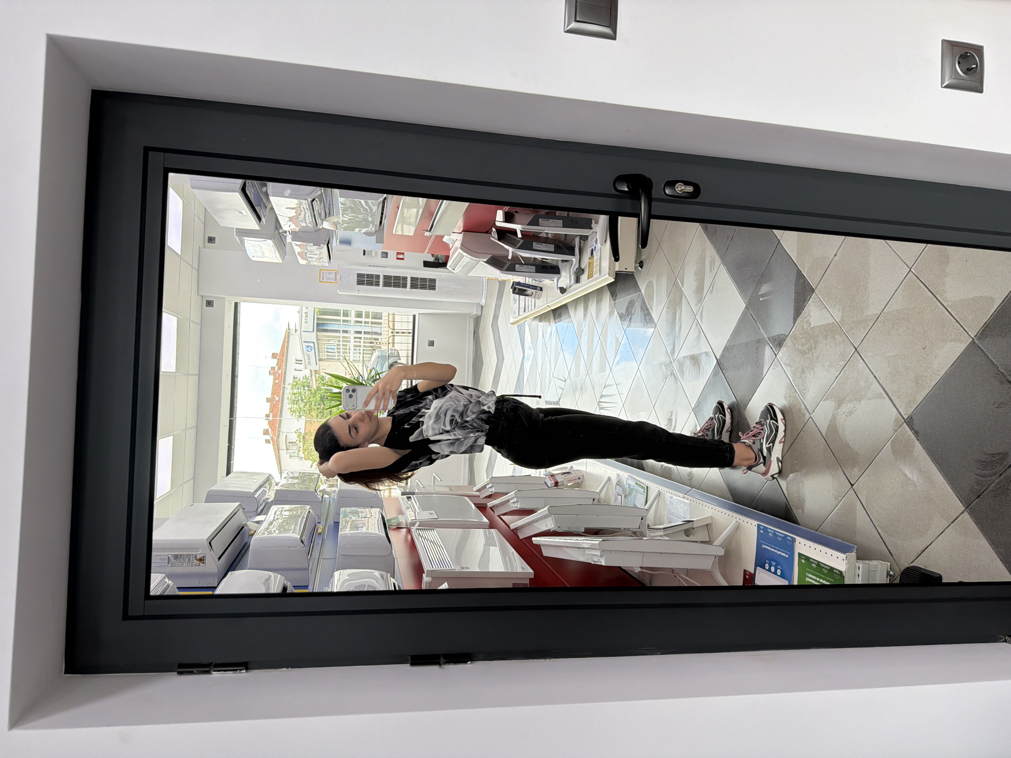 The photograph depicts a young woman, presumably of Eastern European descent, taking a selfie inside what appears to be a retail store, possibly in Gabrovo, Bulgaria.
The woman is dressed in a black and white patterned t-shirt and black pants, along with running shoes. She is holding her phone up and positioned to capture her reflection in what seems to be a large window display, giving the impression she is posing for the camera. Her head is resting on her hands, creating a relaxed pose.
The backdrop of the image is the interior of the store. Shelves are visible, stocked with what appear to be air conditioners and other household appliances. The floor has a diamond pattern design, with alternating light and dark tiles. Outside the storefront, a glimpse of a street scene can be seen, suggesting the store is located in an urban environment.
The photograph is taken from an elevated position, looking down into the window display. The overall tone is casual.