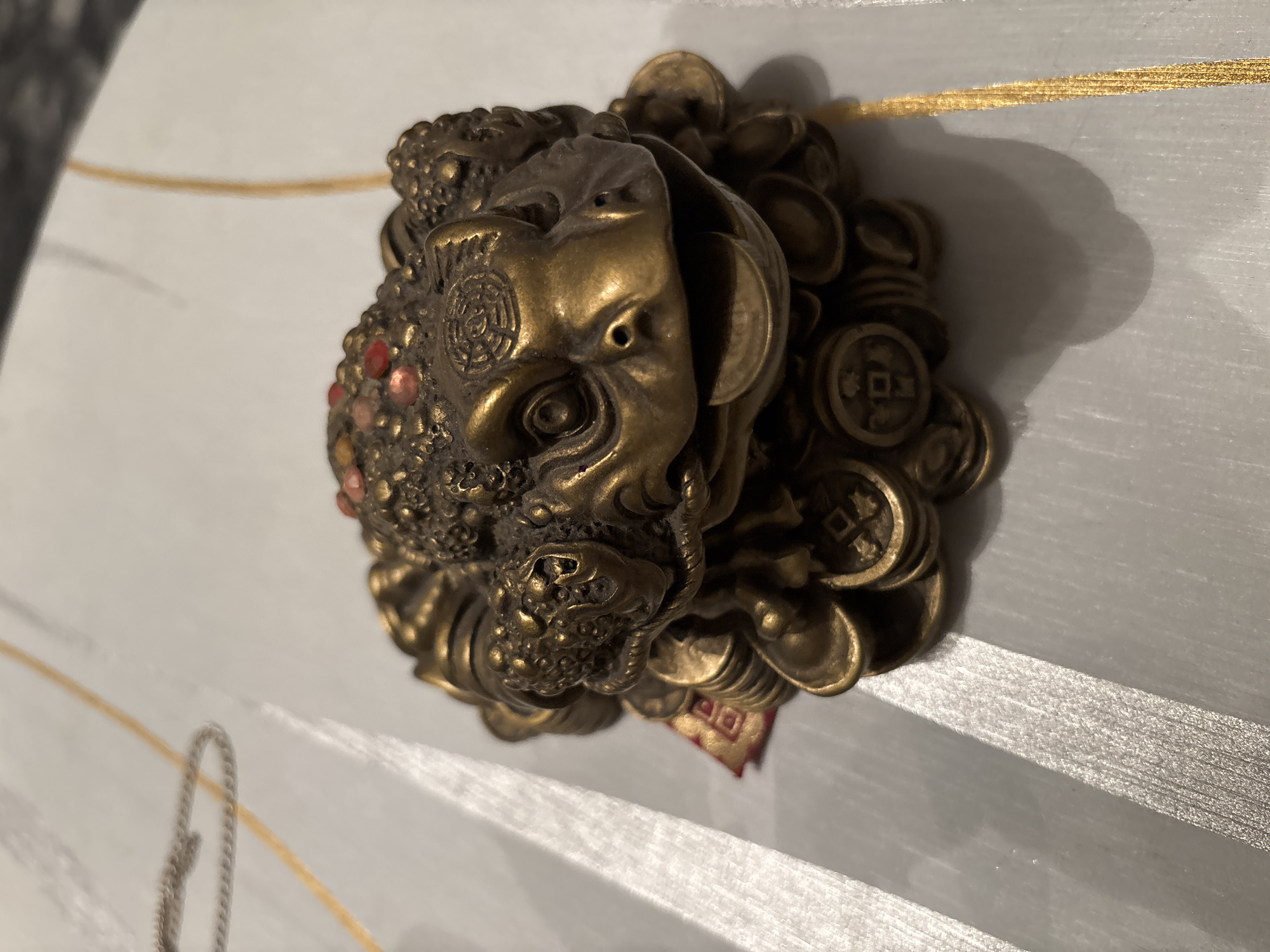 A bronze figurine of a three-legged toad with a coin in its mouth sits on a striped surface with gold and silver lines. The toad is decorated with small red and yellow stones on its back. A portion of a red ribbon with a square pattern is visible beneath the toad. The background is a light-colored, textured surface with visible shadows. The overall scene is an indoor still life. The image was captured in Chernogorsk, Russia.
