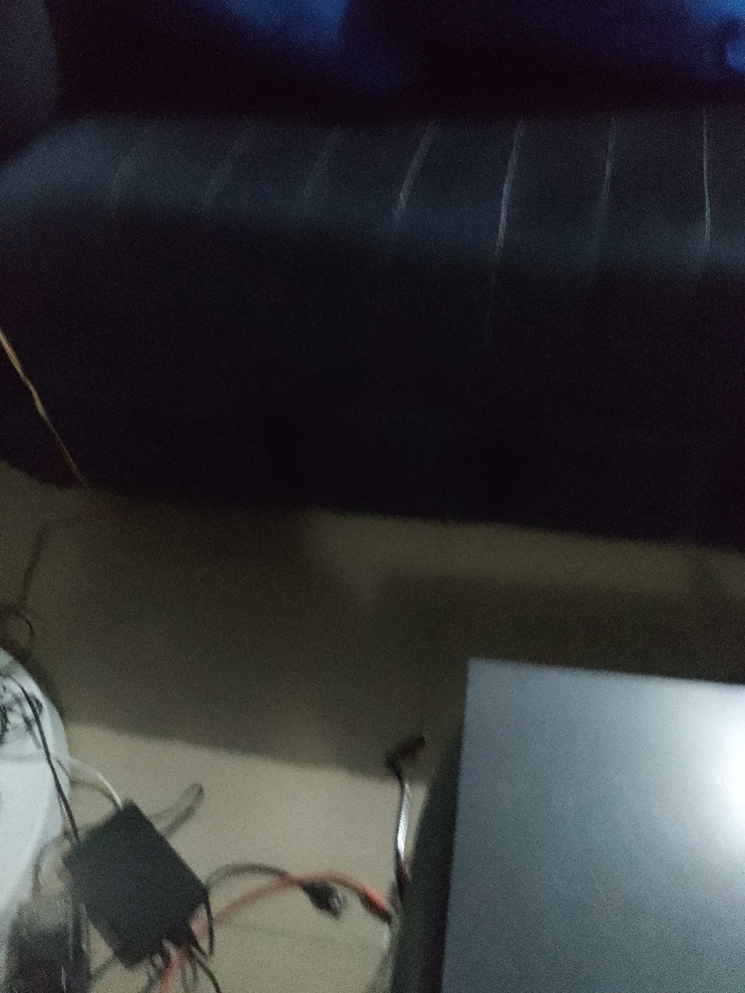 An indoor scene shows a black sofa in the upper half of the image. In the lower half of the image, a black electronic device with wires is on the floor, along with other cables. To the right of the electronic device is a gray, flat object, possibly a monitor or a box. The overall lighting is dim and grainy.