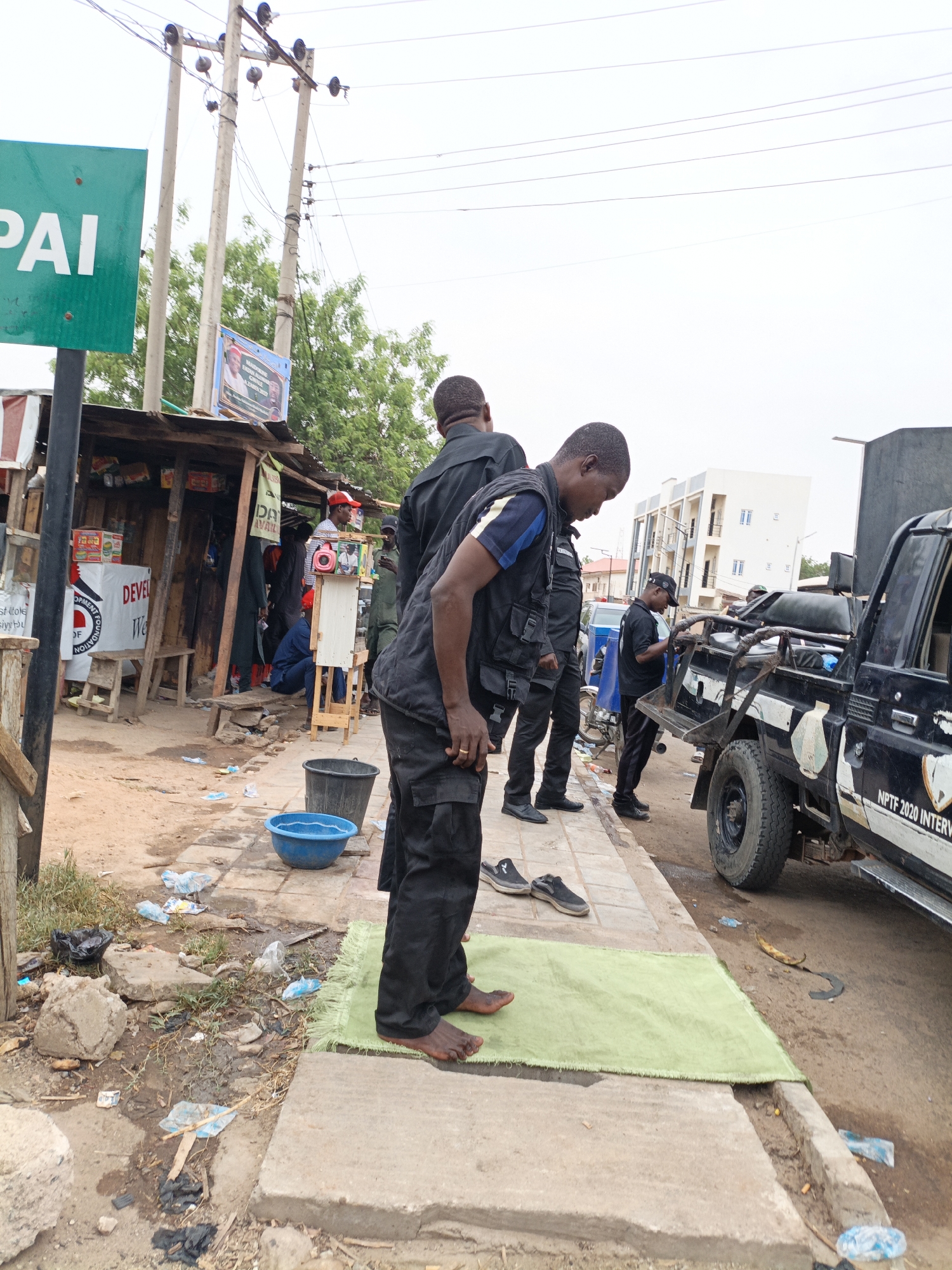 In Tudun Wada, Nigeria, on a day with overcast skies, several law enforcement officers are present on a street beside a parked black pickup truck labeled "NPTF 2020 INTERV." One officer is kneeling barefoot on a small green mat, appearing to be praying. His shoes are removed nearby. Two other officers stand behind him. The area is littered with trash, including plastic bottles and paper. A small roadside shop is visible in the background, along with power lines and other indistinct figures. A green street sign with the letters "PAI" is also visible.