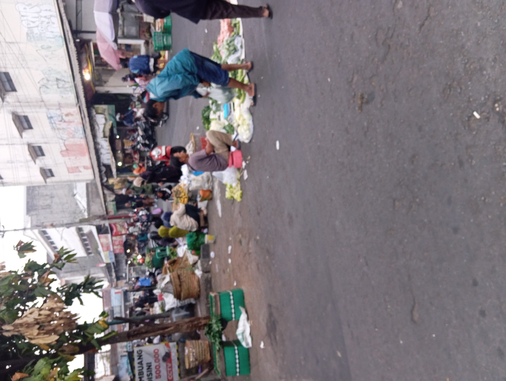 The image captures a bustling street market scene in Yogyakarta, Indonesia. In the foreground, a person in a teal jacket is bending over, handling produce, while another individual in a brown shirt and tan pants sits on a red stool. The ground is paved with asphalt and appears wet in some areas. Various vendors' stalls are visible, displaying a variety of fruits and vegetables. Baskets and crates are scattered around, filled with goods. In the background, there are buildings with a commercial appearance, some showing signs of graffiti. Motorbikes are parked and in motion, indicating the presence of transportation. The overall atmosphere suggests a typical day in a busy Indonesian marketplace.