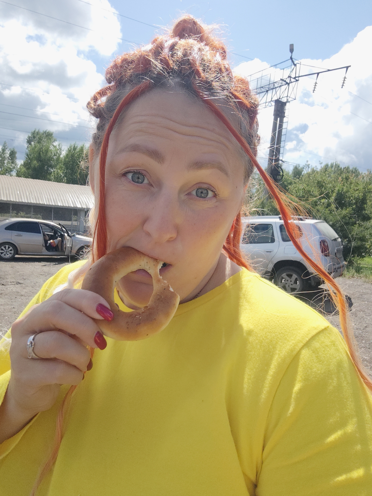 A woman with orange dreadlocks, wearing a bright yellow long-sleeved shirt, is shown from the chest up. She is holding a round, baked good, possibly a bagel or a roll, to her mouth and appears to be biting into it. She is wearing a silver ring on her left ring finger, and her fingernails are painted a dark pink. Her eyes are blue and she is looking directly at the camera. The background includes a silver car with its door open, a white SUV, a building with a corrugated metal roof, trees, and a power line tower against a cloudy blue sky. The ground appears to be gravel or dirt. The scene is outdoors, likely in a rural or industrial area, possibly on the outskirts of Kambarka, Russia, given the context of the provided crops.
