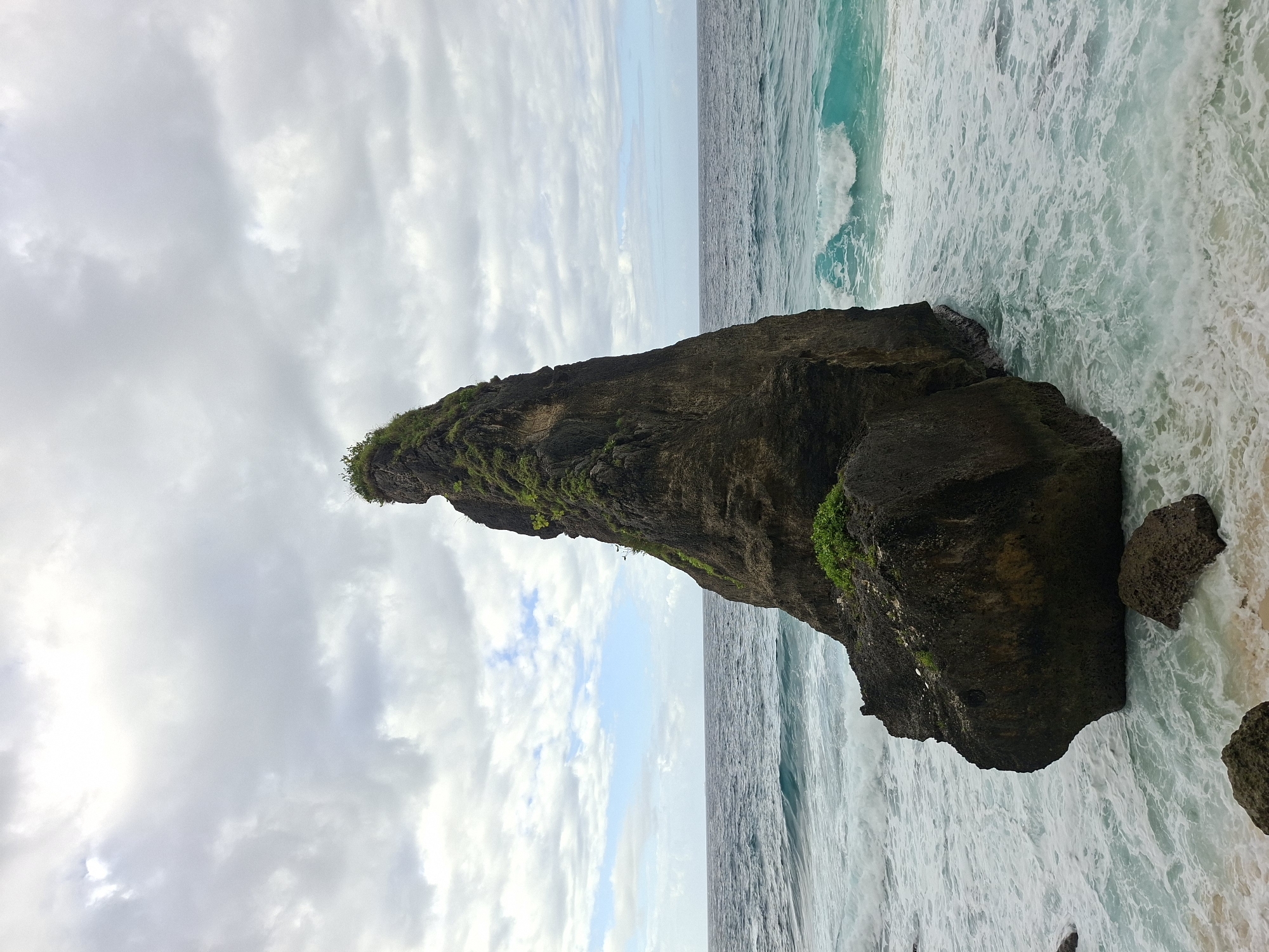 The image depicts a large, dark-colored rock formation partially submerged in the ocean, with vegetation growing on its peak. The rock appears rugged and textured, contrasting with the surrounding turquoise waters and white foamy waves. To the left of the rock, the sky is filled with gray, cloudy skies with glimpses of blue sky. The horizon line is visible in the middle of the photo. The location is Nusa Penida, Indonesia.
