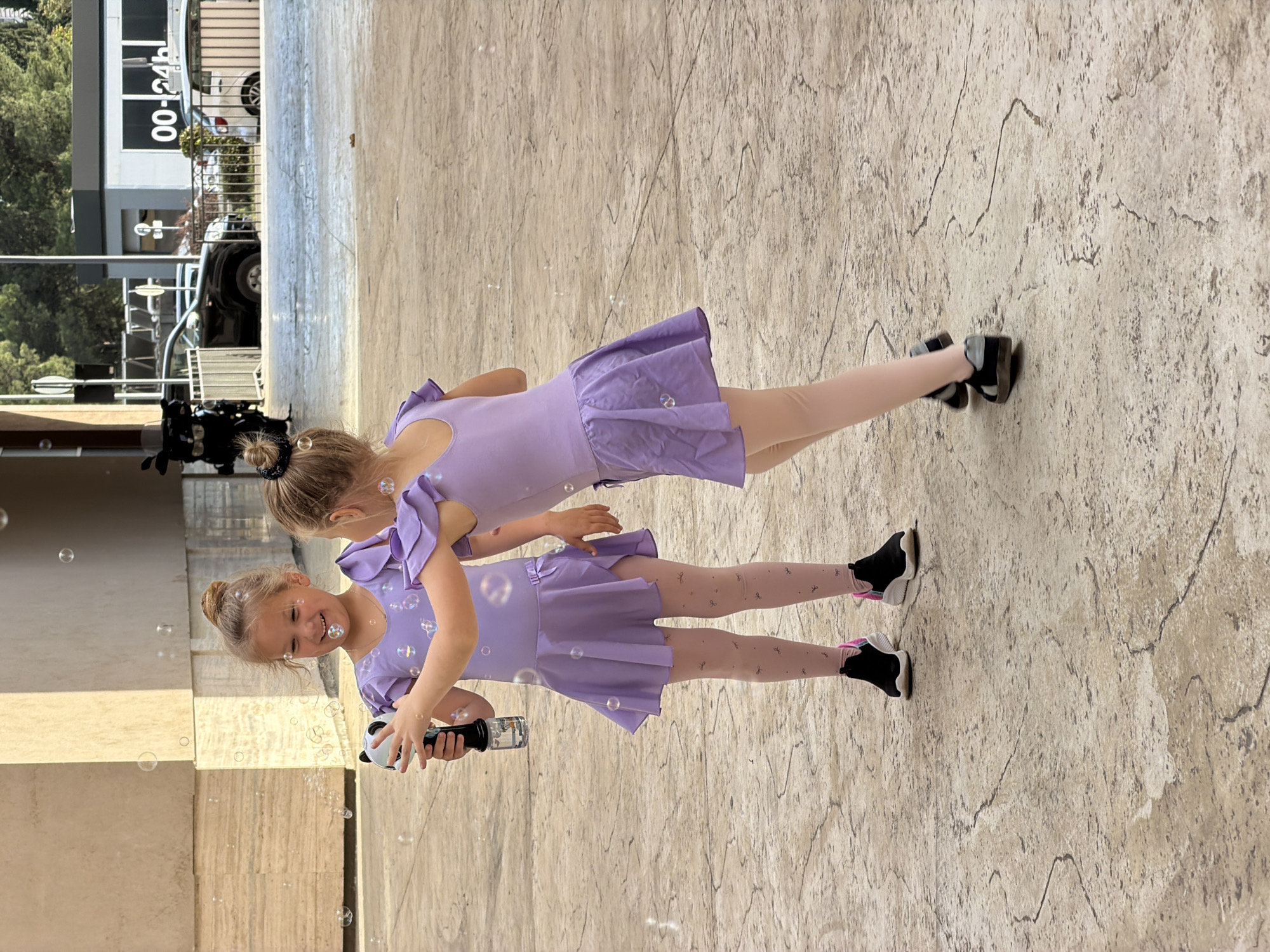 Two young girls are captured outdoors, likely playing in Budva, Montenegro. Both girls have light-colored hair styled in buns and are dressed in matching light purple ballet-style leotards or short dresses with ruffled sleeves and flared skirts. They wear light-colored tights adorned with small bow patterns and black sneakers; one pair has pink accents, and the other has white.
The girl in the foreground, smiling broadly and appearing joyful, holds a black and white bubble gun, possibly shaped like a panda, from which numerous translucent soap bubbles are emerging. The second girl stands slightly behind her, looking towards the first, and appears engaged in the bubble play.
They are positioned on a light-colored, textured pavement with dark, irregular lines resembling cracked stone. In the background, a modern building with glass sections is visible, along with a black vehicle and a metal railing. White text "00-24h" can be seen on the building, indicating operating hours. The scene is brightly lit, suggesting a sunny daytime. The overall atmosphere is cheerful and playful.